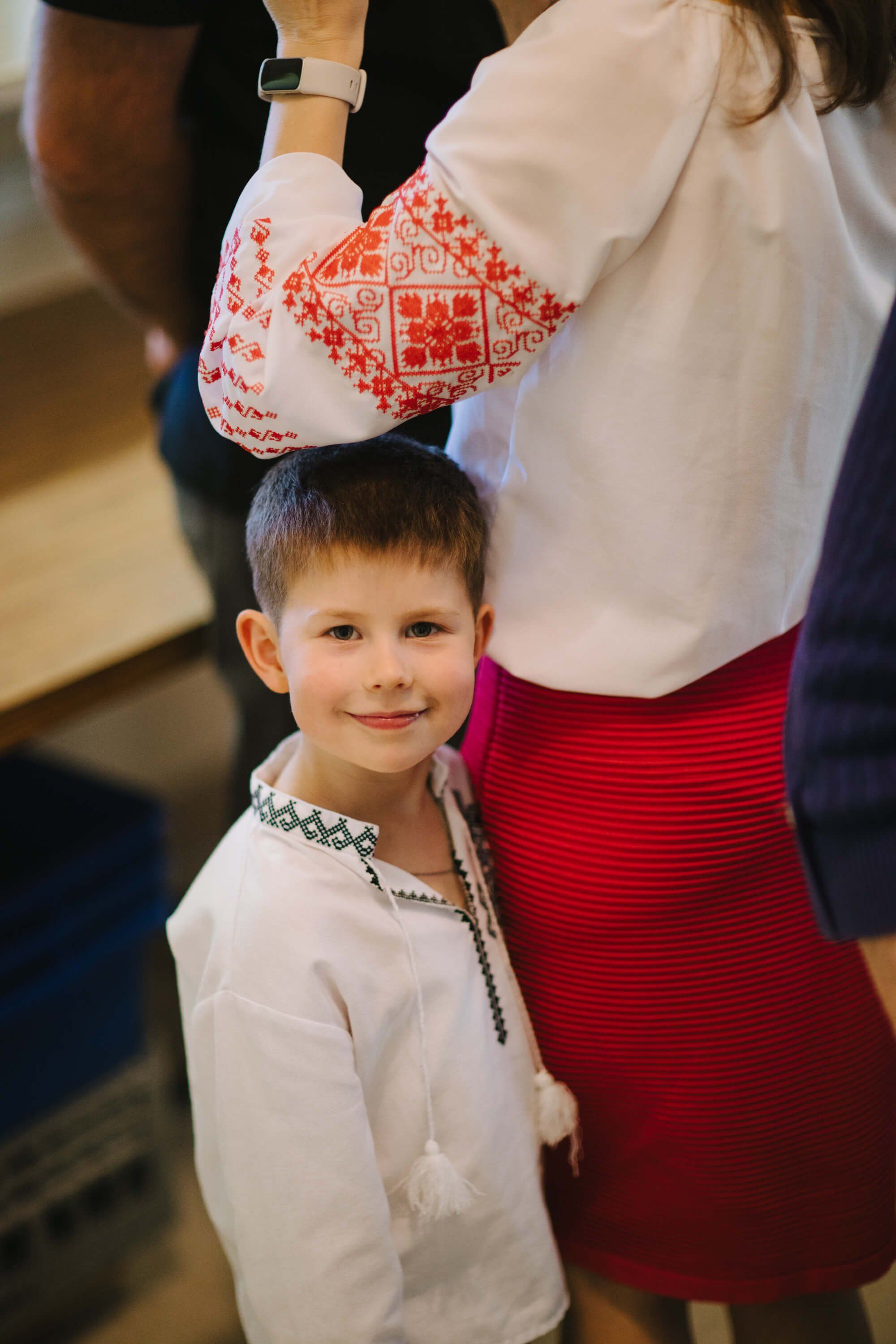 Young boy in embroidered white shirt smiles beside woman wearing traditional Ukrainian red skirt and ornate white embroidered vest