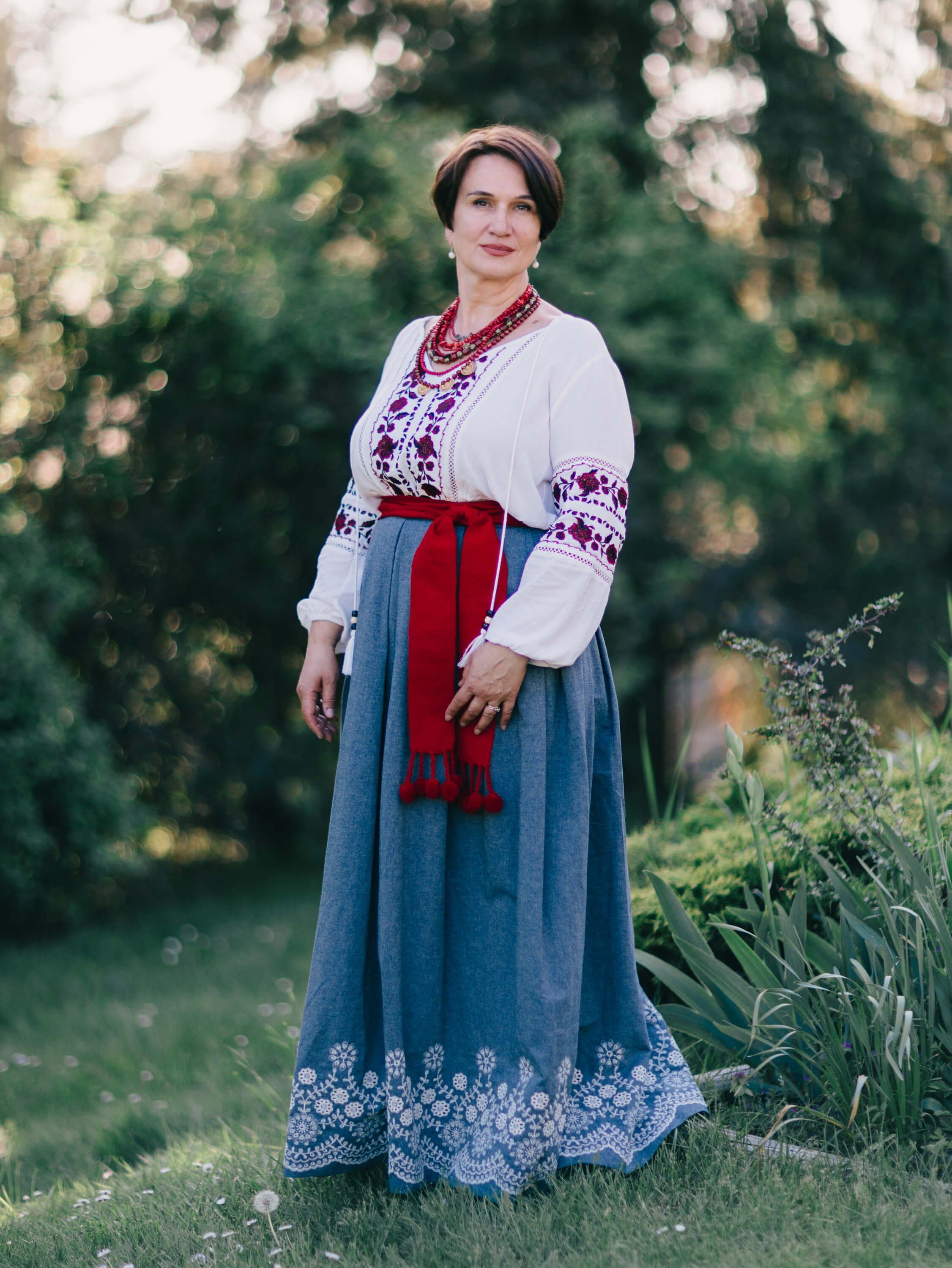 Woman in traditional Ukrainian embroidered dress and red sash stands in golden-hour garden light, serene and poised