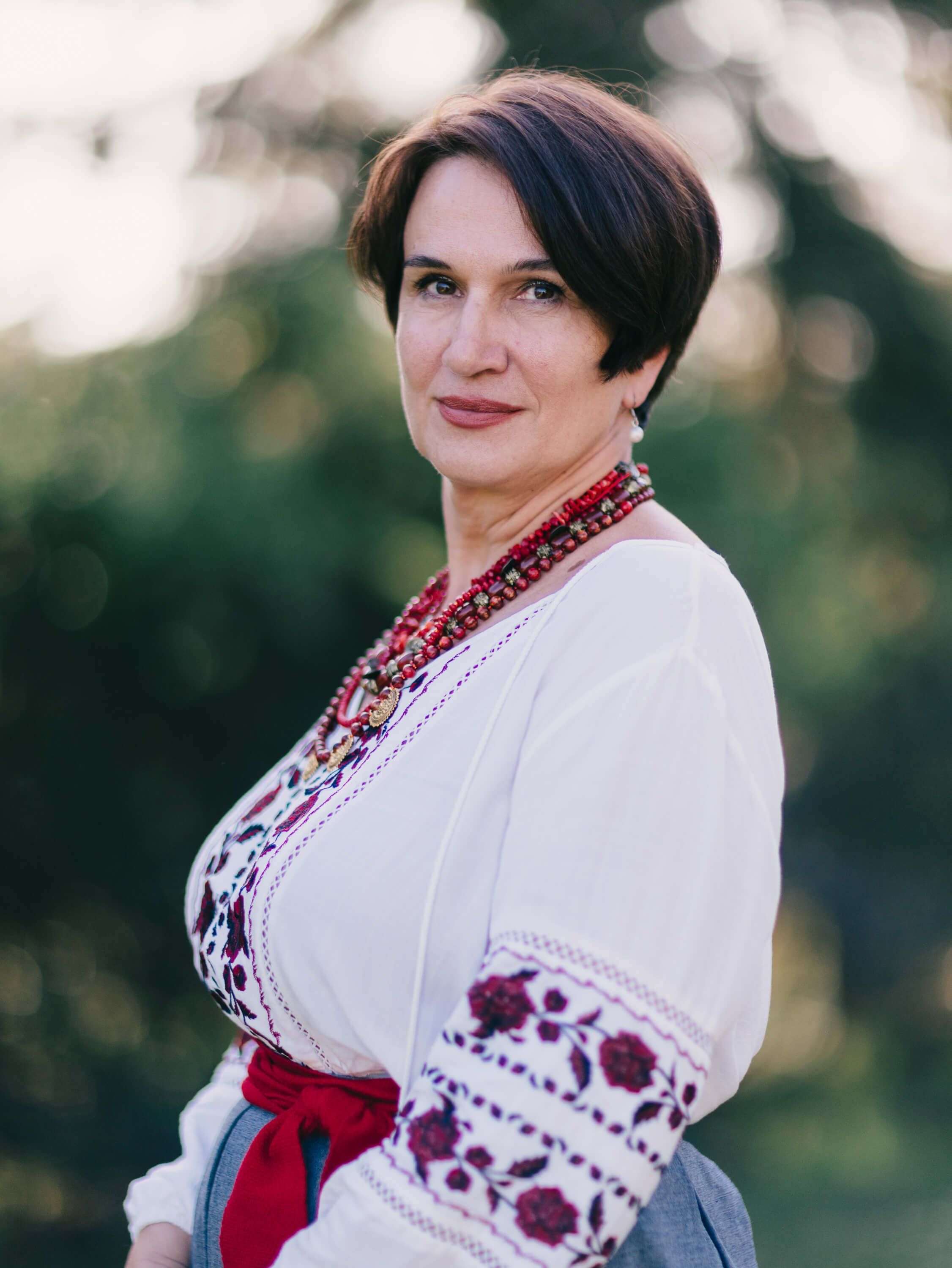 Woman in traditional Ukrainian embroidered white linen blouse with red beading and red sash, posed against soft-focused green background in golden hour light