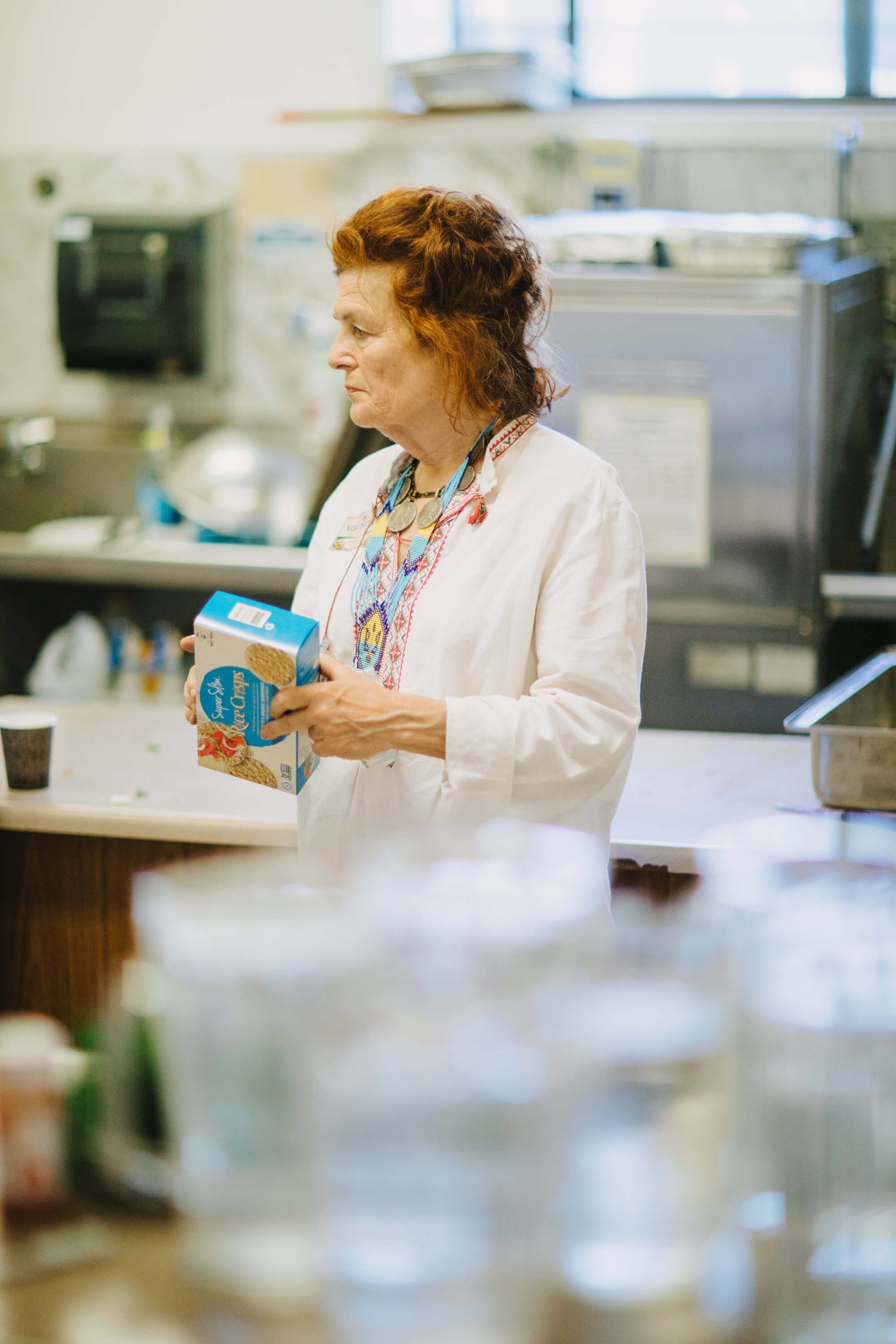 Woman with red hair holding a blue box in a bright kitchen, smiling while speaking at the fundraiser dinner