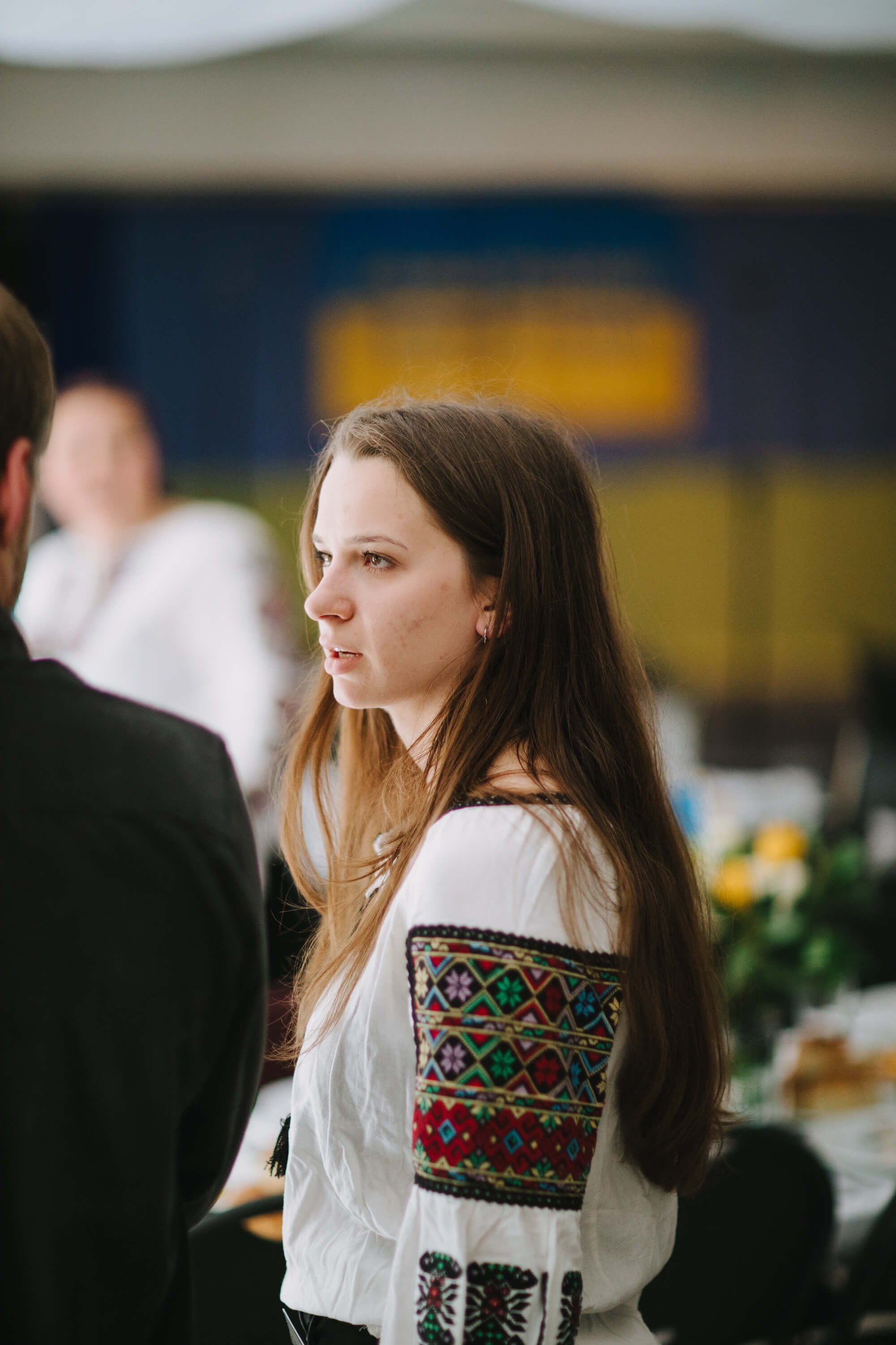 Woman in embroidered white blouse at indoor event, soft natural light, composed expression, blurred yellow and blue background