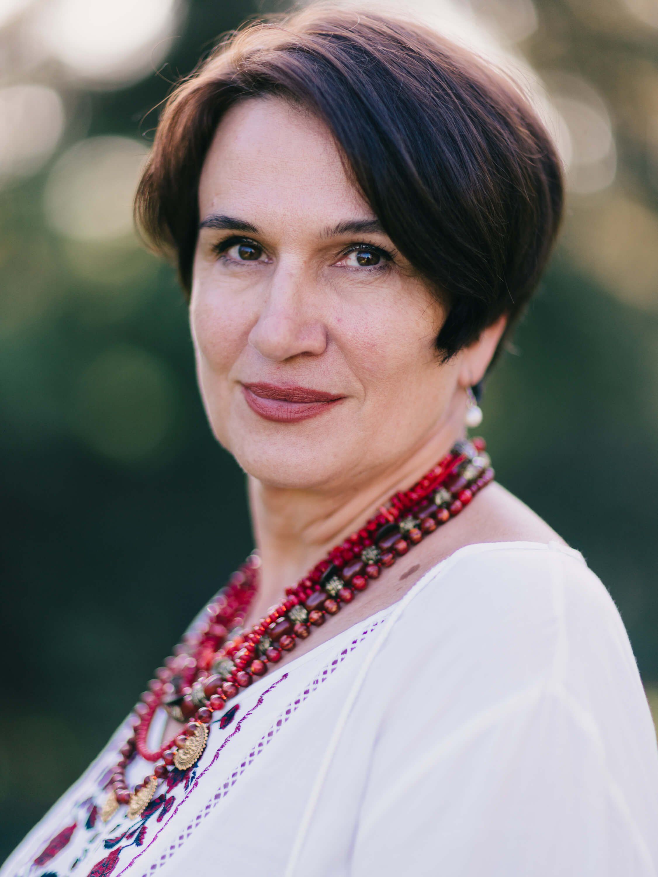 Woman in embroidered white linen shirt and red beaded necklace smiles warmly at camera against soft green bokeh background