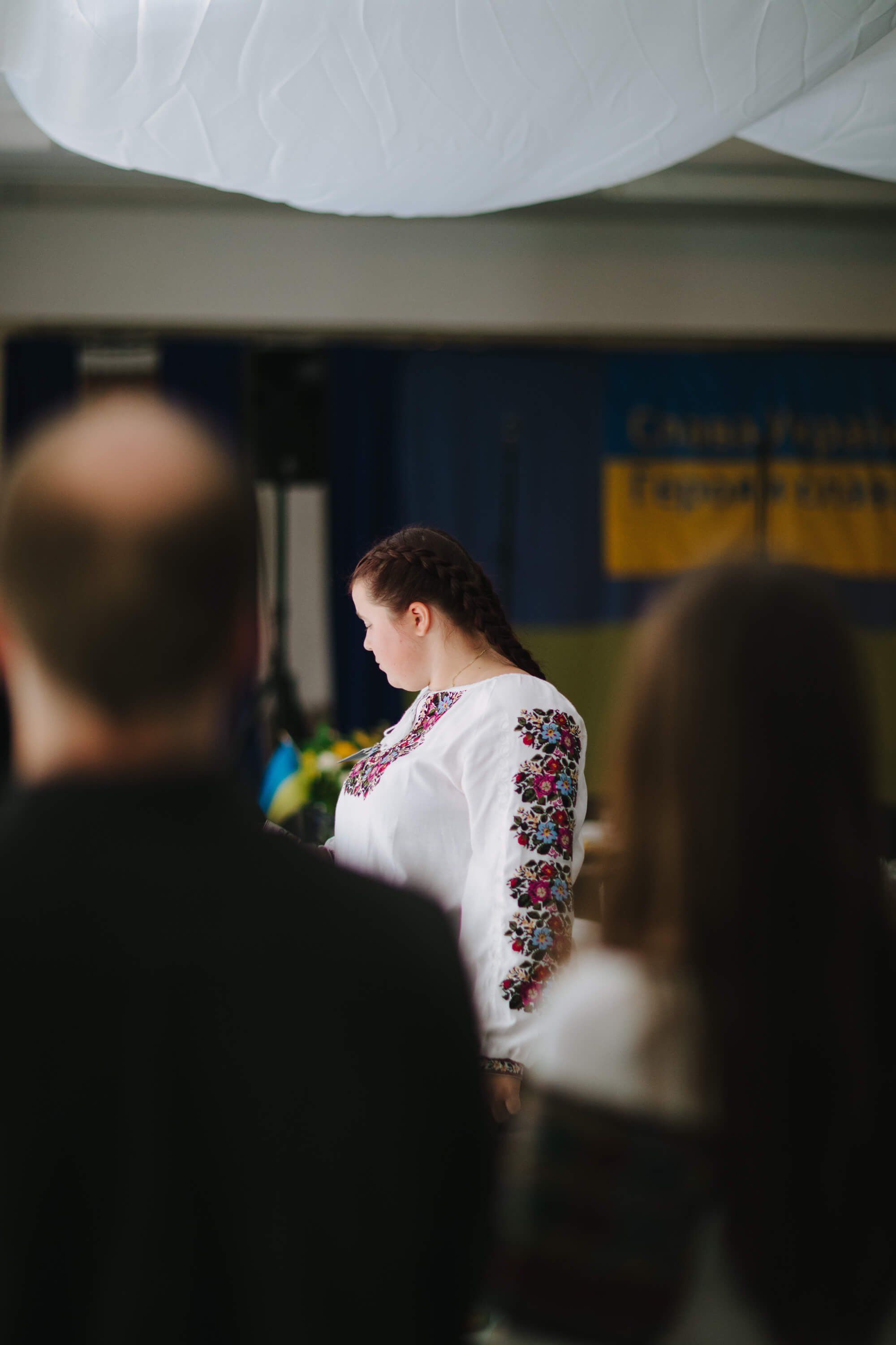 Woman in embroidered white blouse speaks at podium during Ukrainian fundraiser dinner, surrounded by attendees