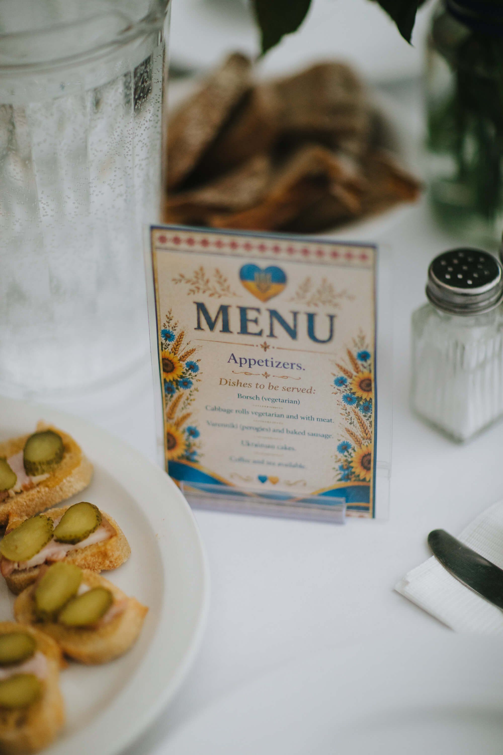 Decorative menu card with blue and gold floral design sits on white tablecloth between appetizer plates and salt shaker at fundraiser dinner