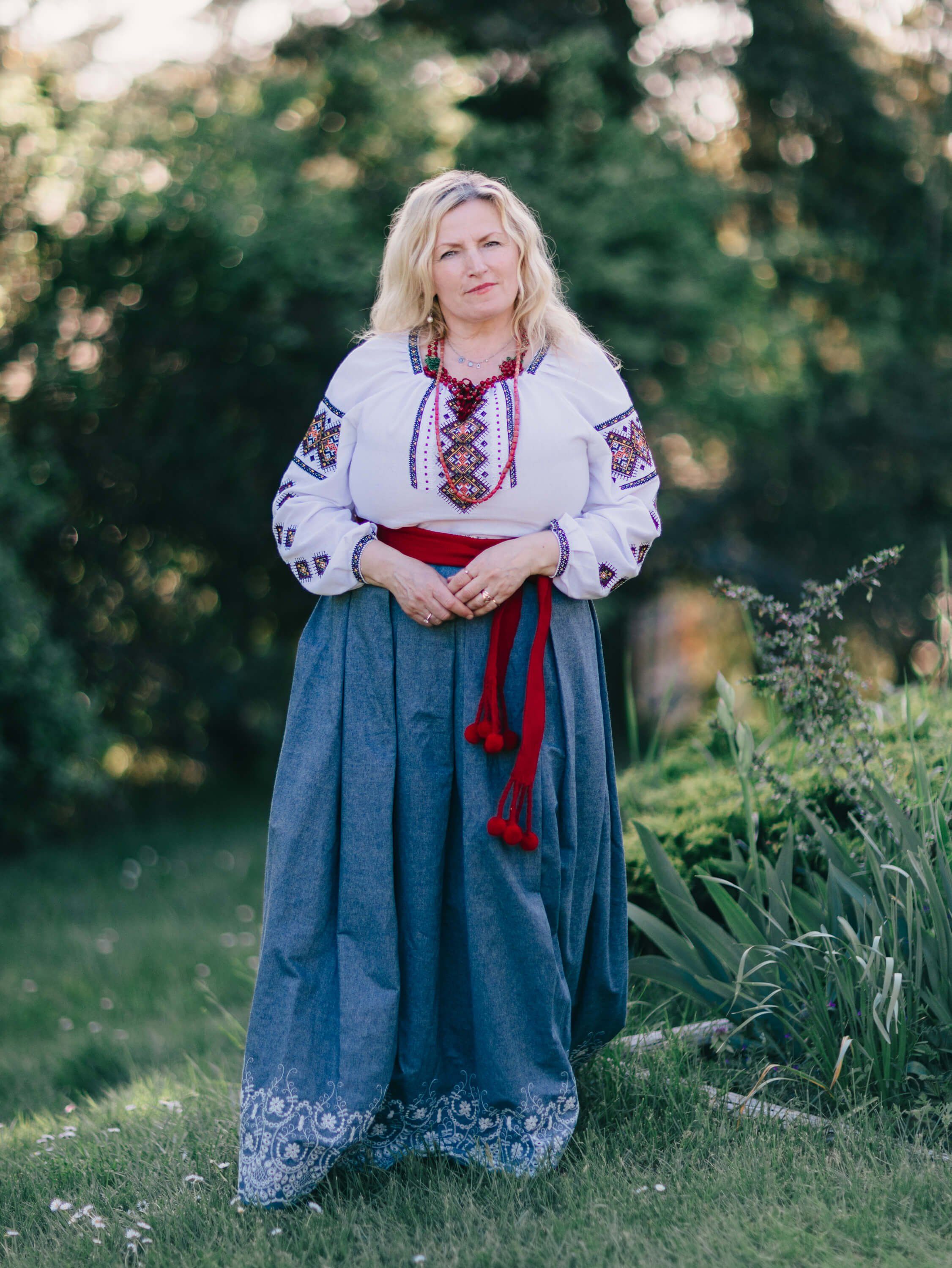 Woman in traditional Ukrainian embroidered blouse and denim skirt stands in golden-hour garden light