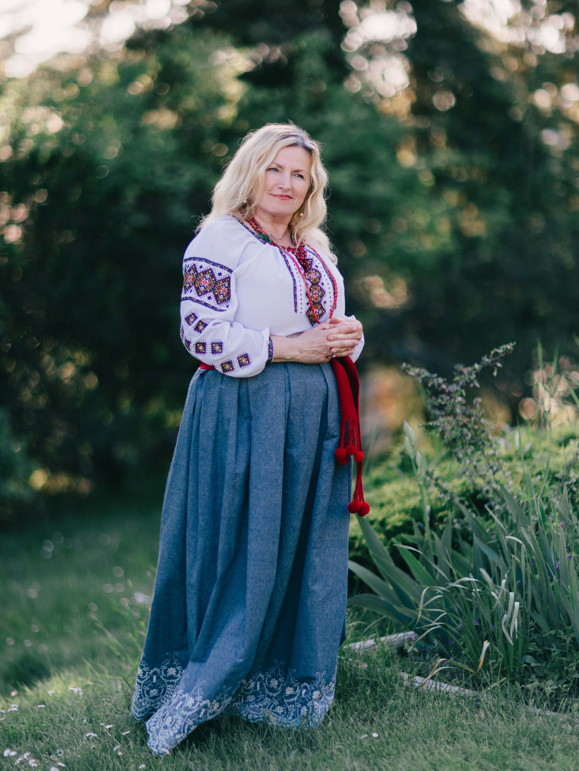 Woman in embroidered white blouse and blue linen skirt stands in golden evening light among garden greenery
