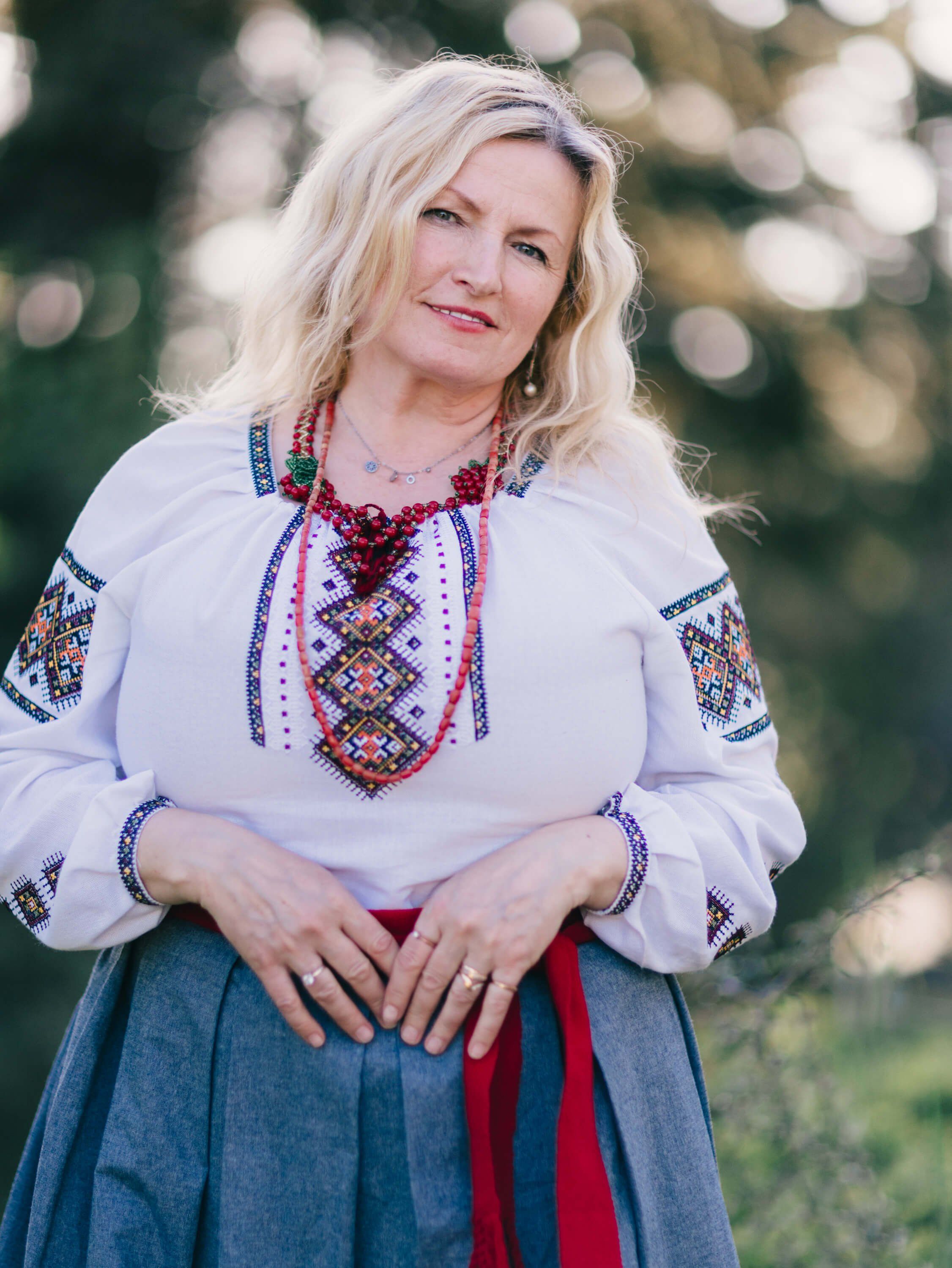 Woman in traditional Ukrainian embroidered white blouse and blue skirt smiles at camera in soft outdoor light