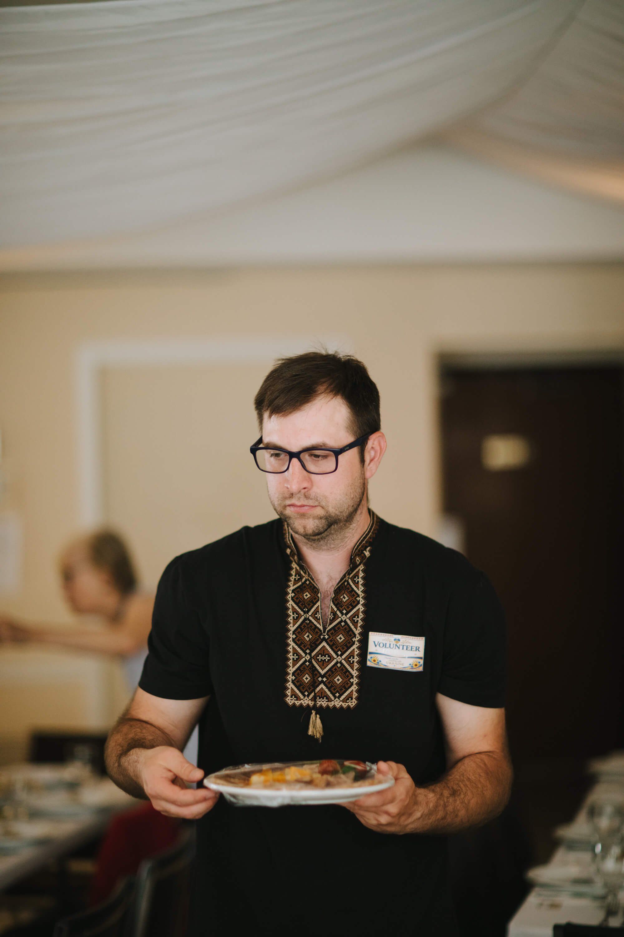Man in glasses holding a plate of food at an indoor fundraiser dinner event