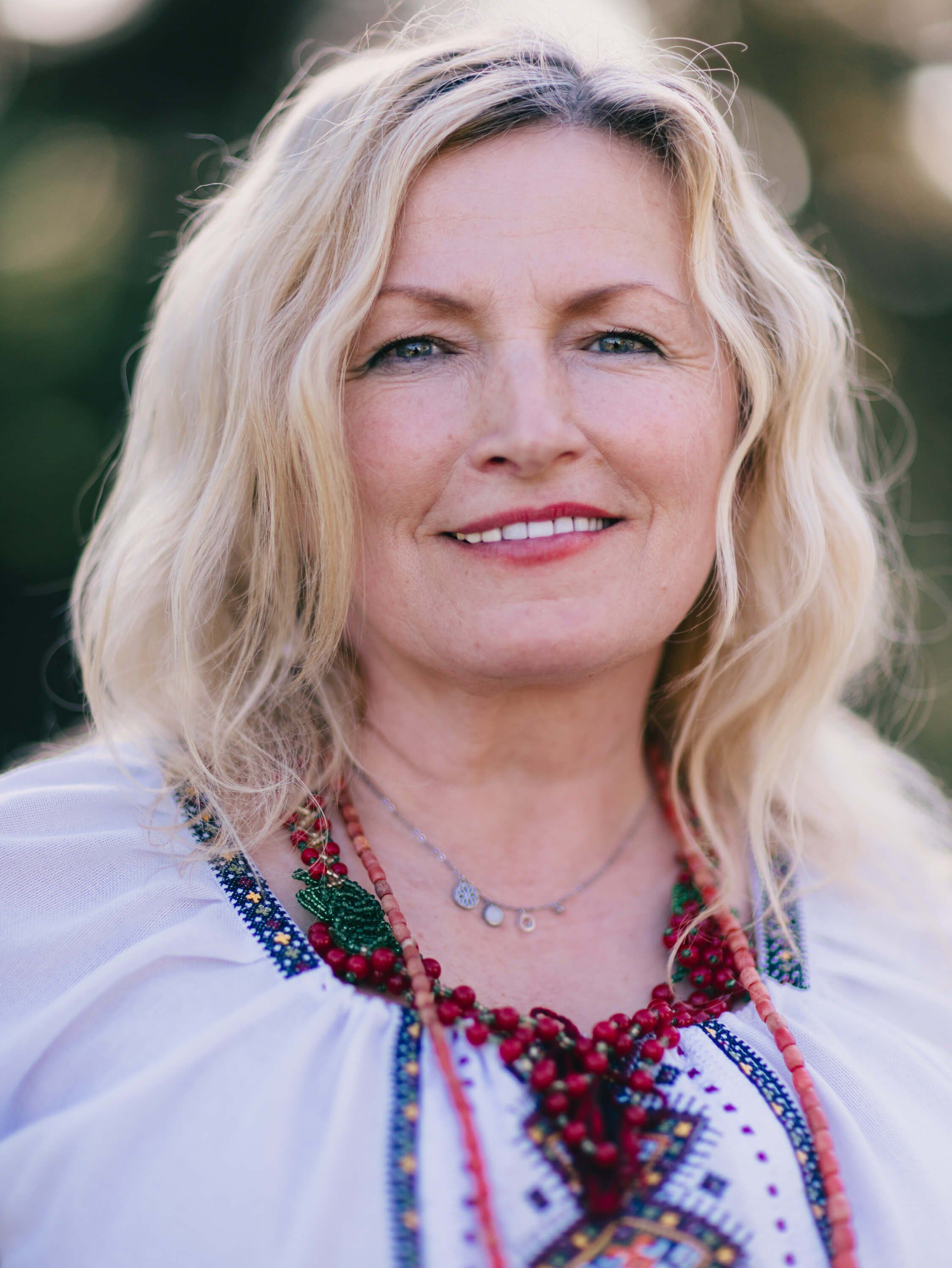 Woman with blonde wavy hair wearing embroidered Ukrainian clothing smiles warmly at the camera outdoors