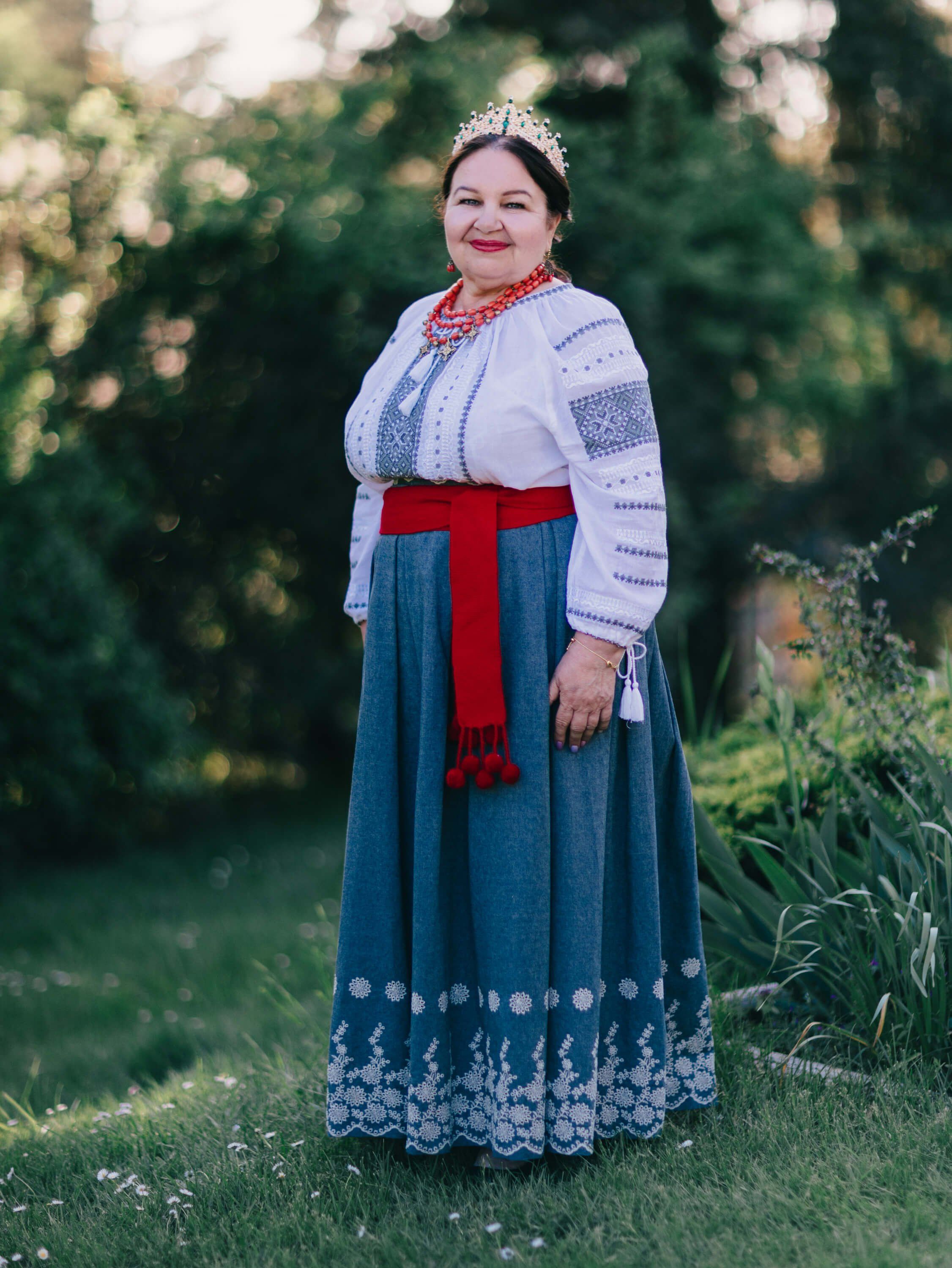 Woman in traditional Ukrainian embroidered white blouse and teal skirt with red sash, tiara, smiling toward camera in golden-hour garden light