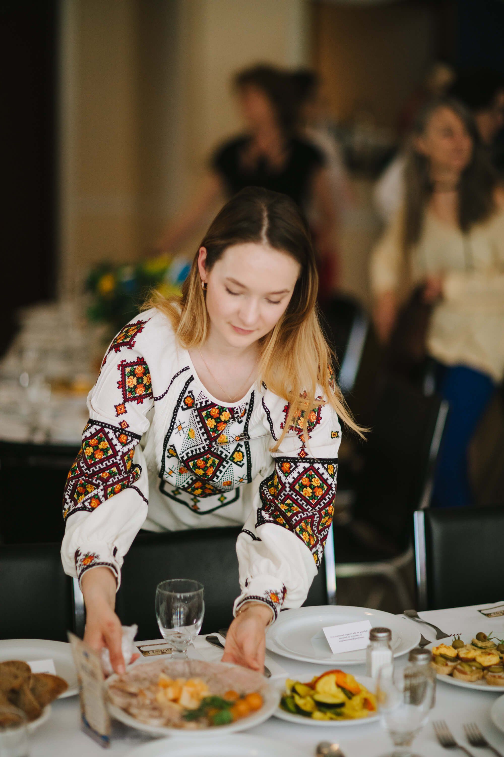 Woman in embroidered white blouse serves herself at a candlelit dinner table surrounded by guests and fresh food