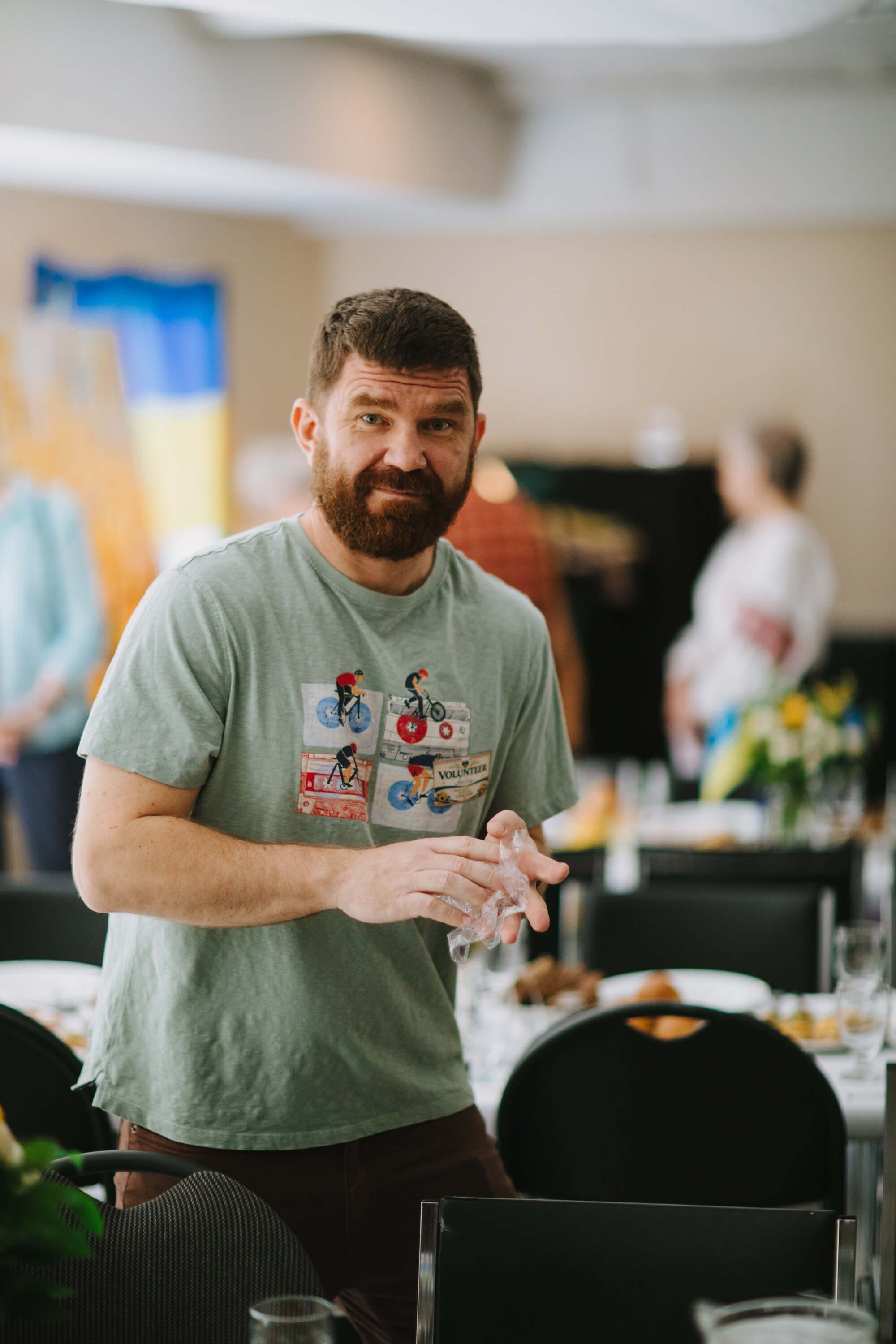 Bearded man in gray graphic tee smiles warmly while standing at a table during an indoor fundraiser event