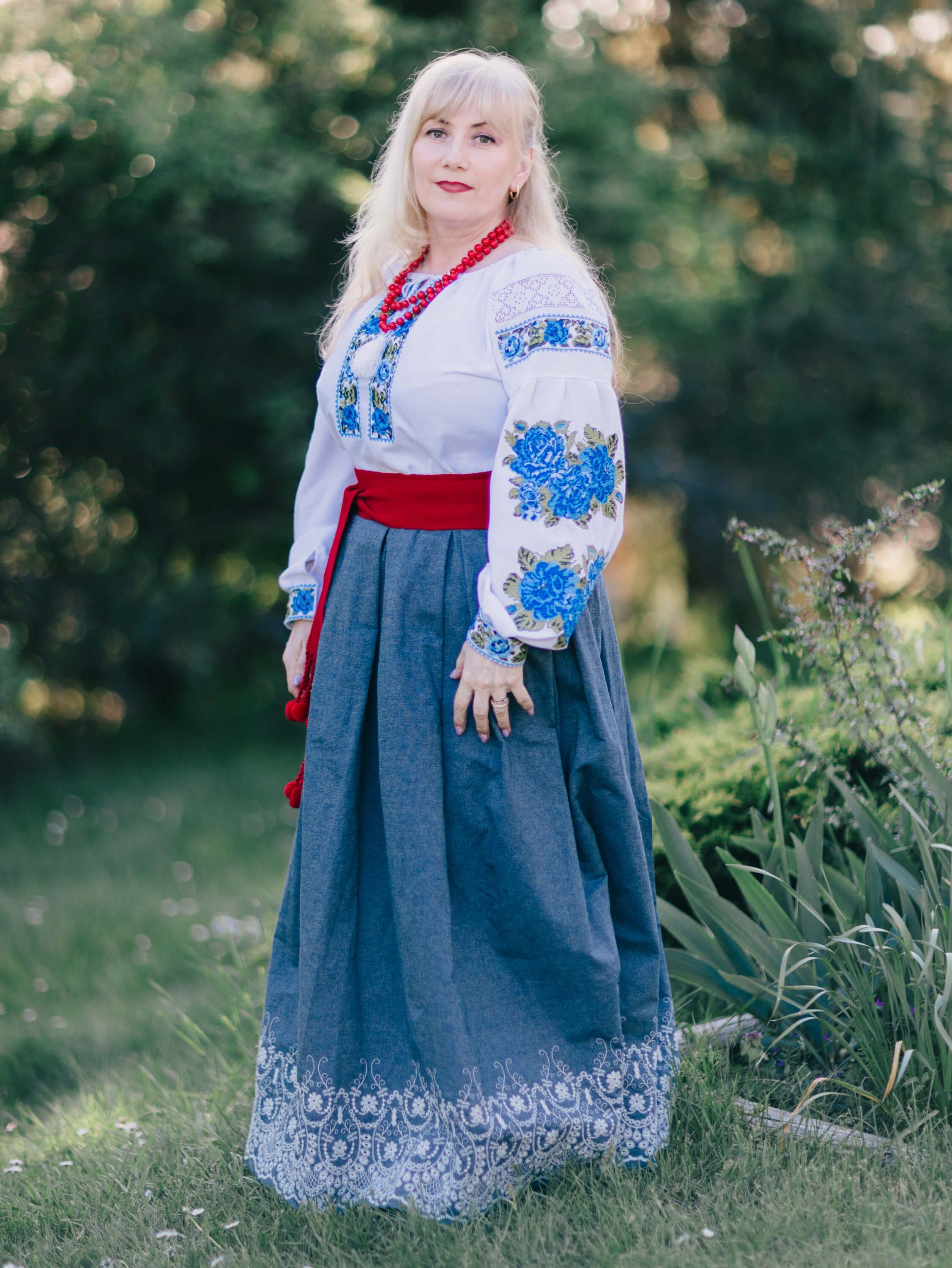 Woman in embroidered Ukrainian white linen shirt and blue skirt with red sash stands in golden evening light among garden greenery