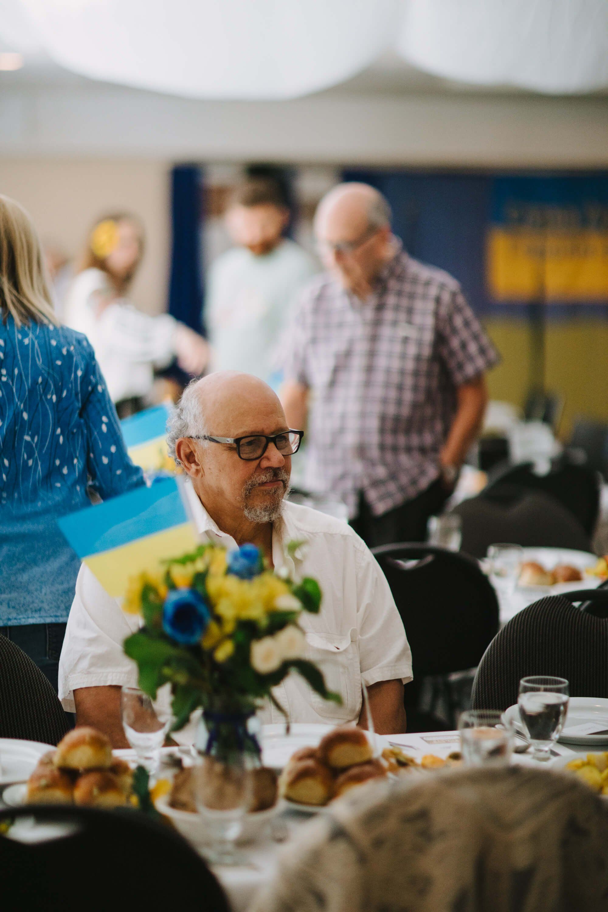Elderly man in glasses and white shirt sits at a dinner table with sunflowers, others mingling in the blurred background of an indoor fundraiser event