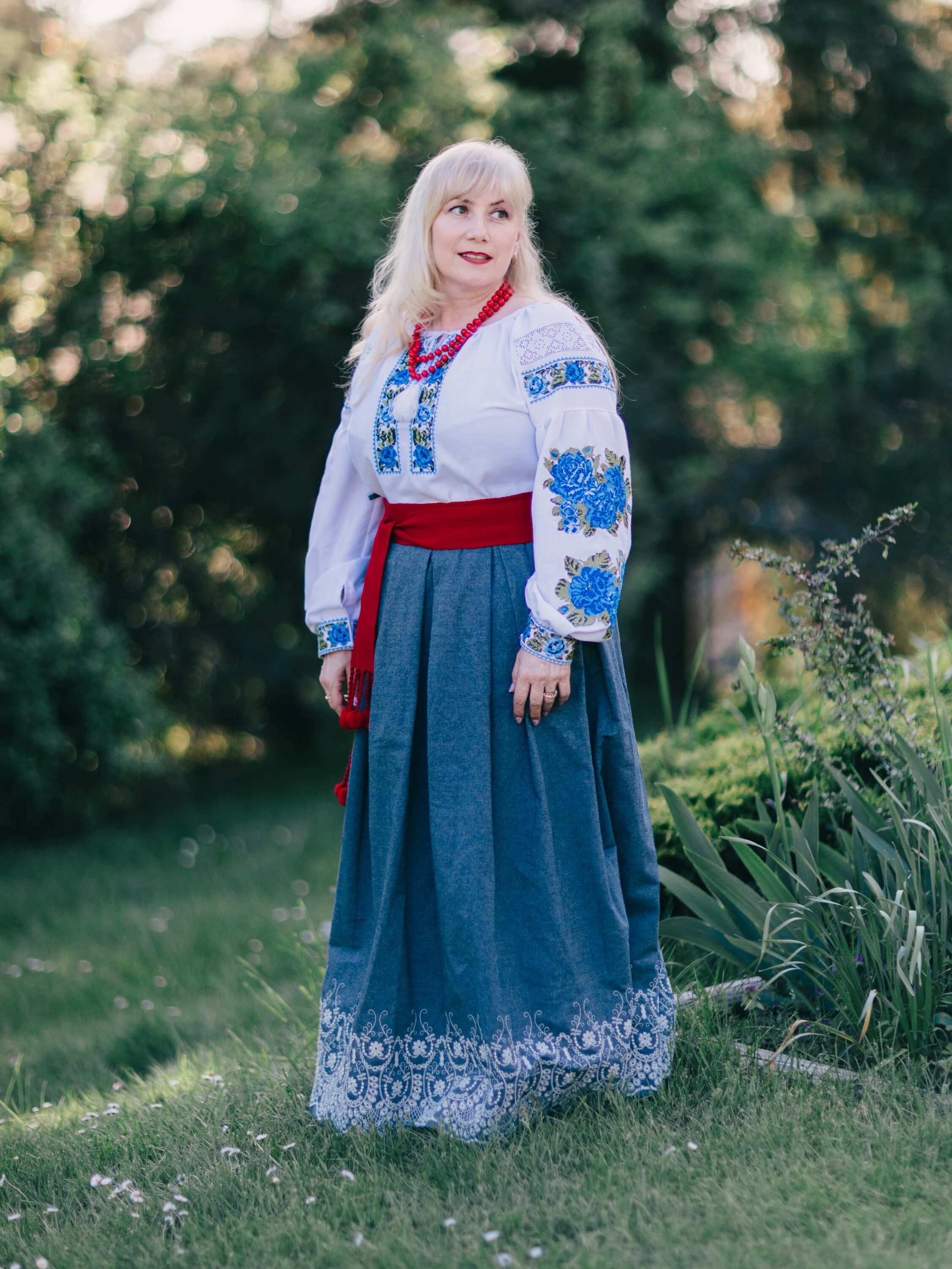 Woman in traditional Ukrainian embroidered blouse and blue skirt stands in golden-hour garden light, looking upward thoughtfully