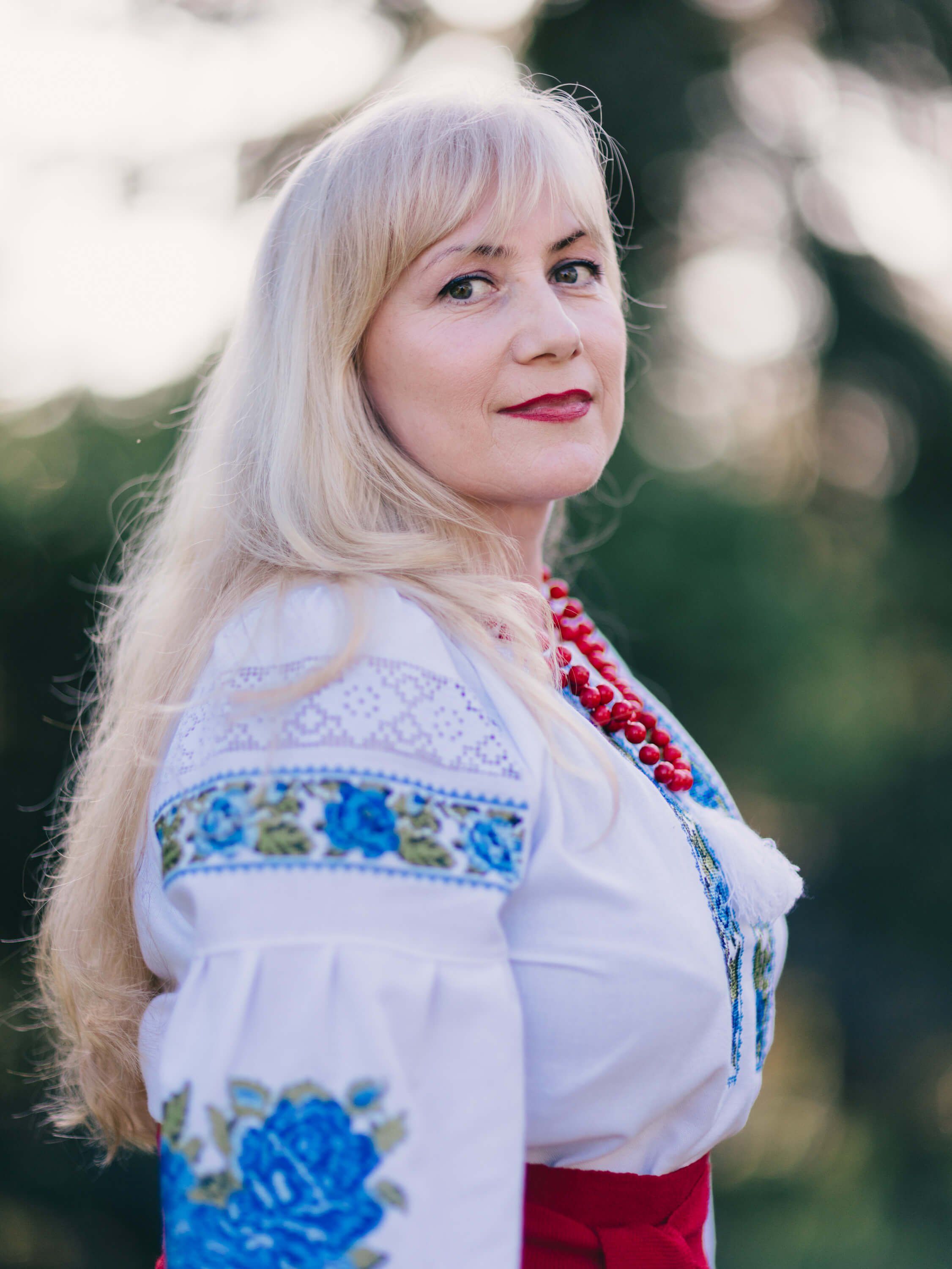 Woman in embroidered white blouse and red skirt with beaded necklace, soft evening light filtering through trees behind her