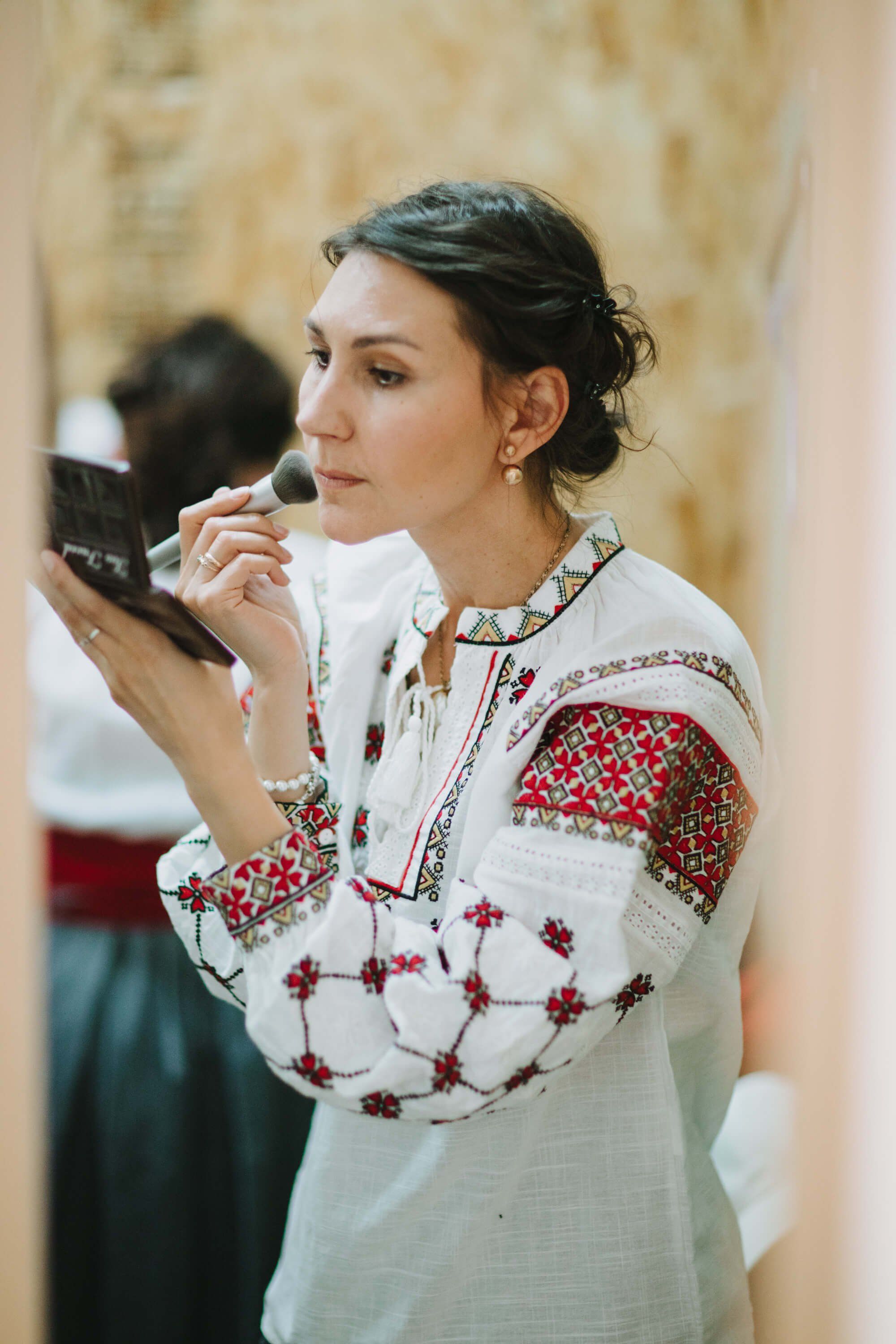 Woman in embroidered white vyshyvanka applies lipstick while holding a compact mirror, warm indoor lighting