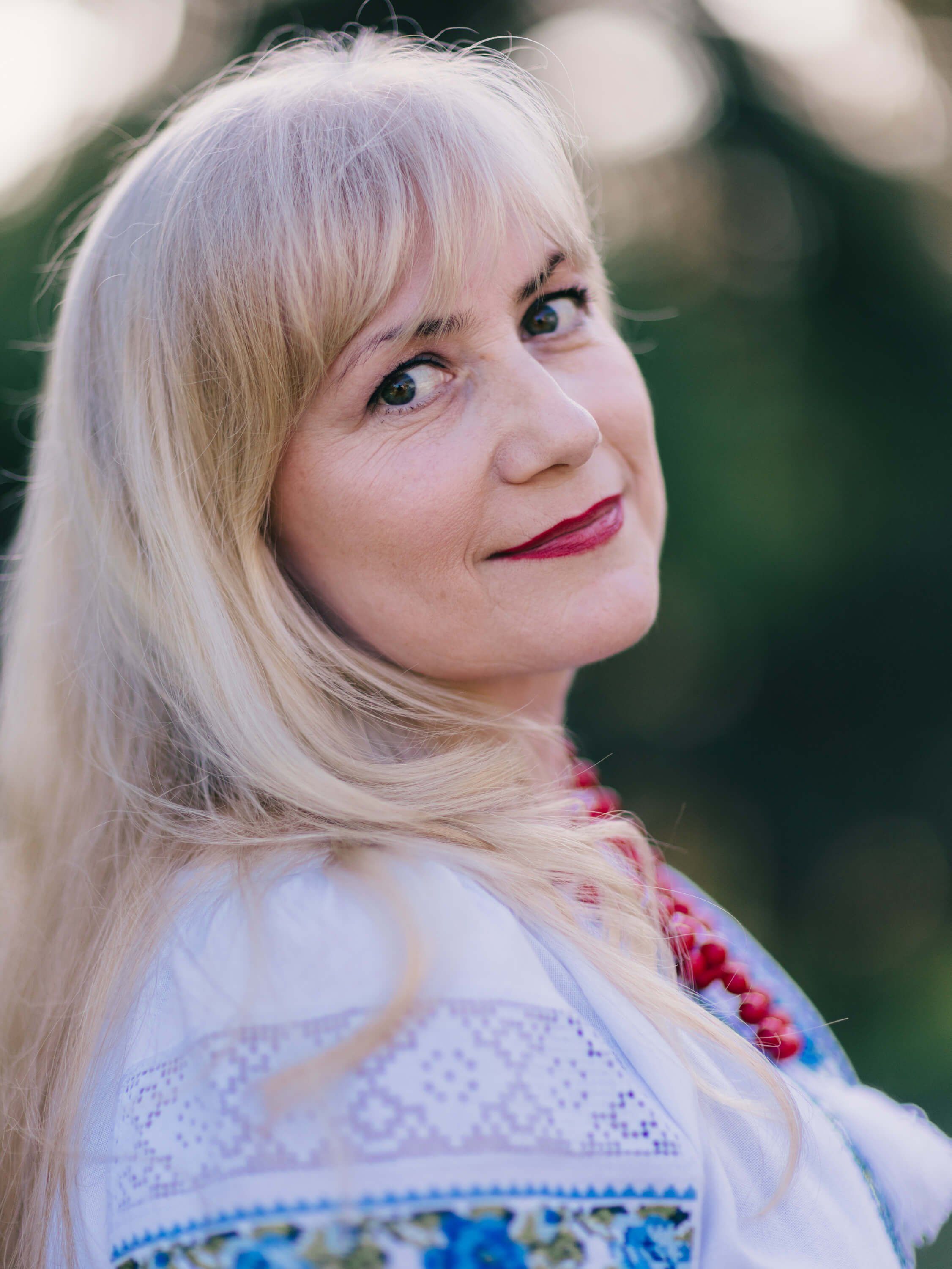 Woman with platinum blonde hair and magenta lipstick smiles warmly at camera, wearing embroidered blue and white garment against soft evening light