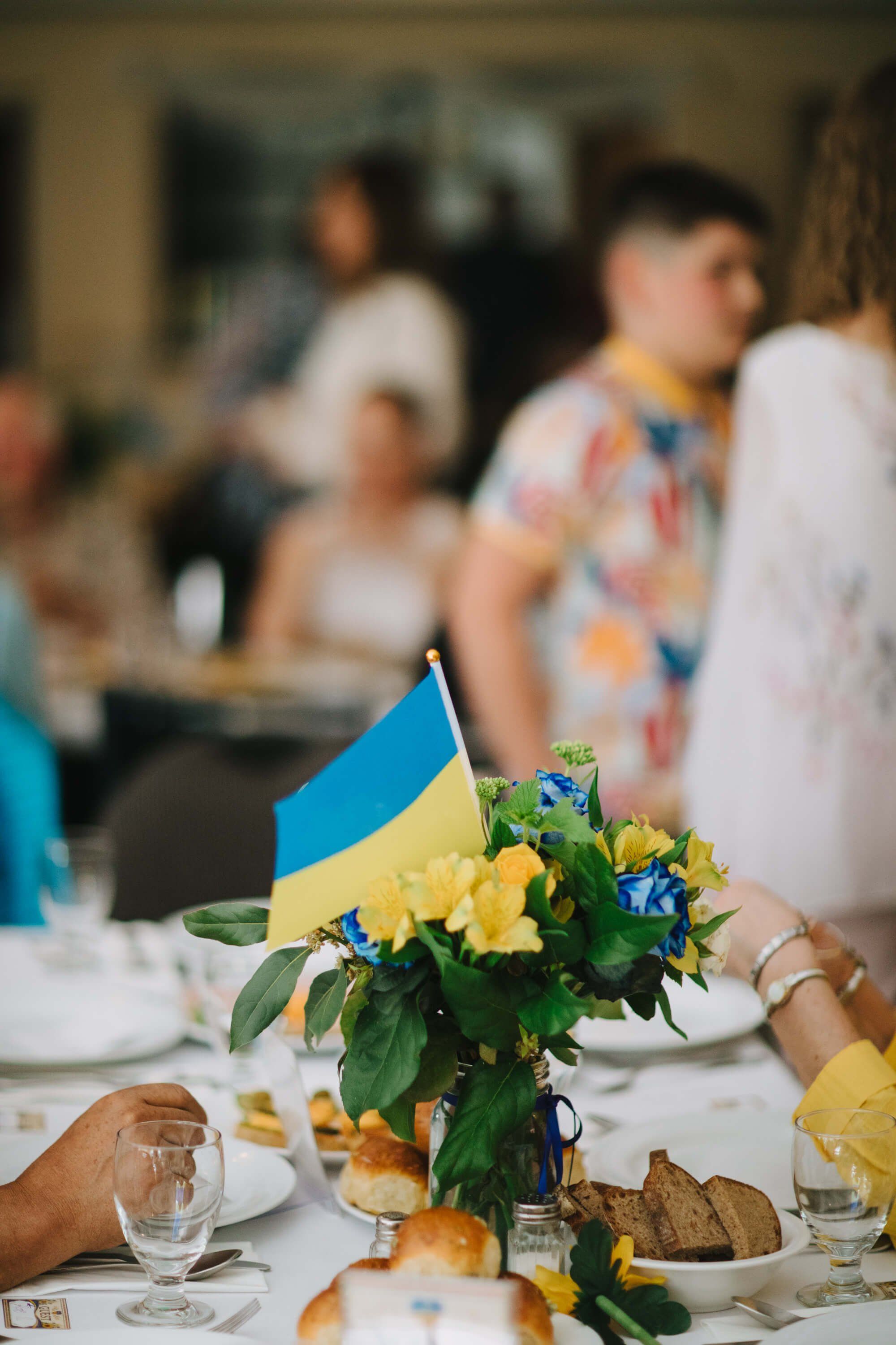Table centerpiece with Ukrainian flag, yellow flowers, and blue blooms beside place settings as dinner guests gather in soft background light