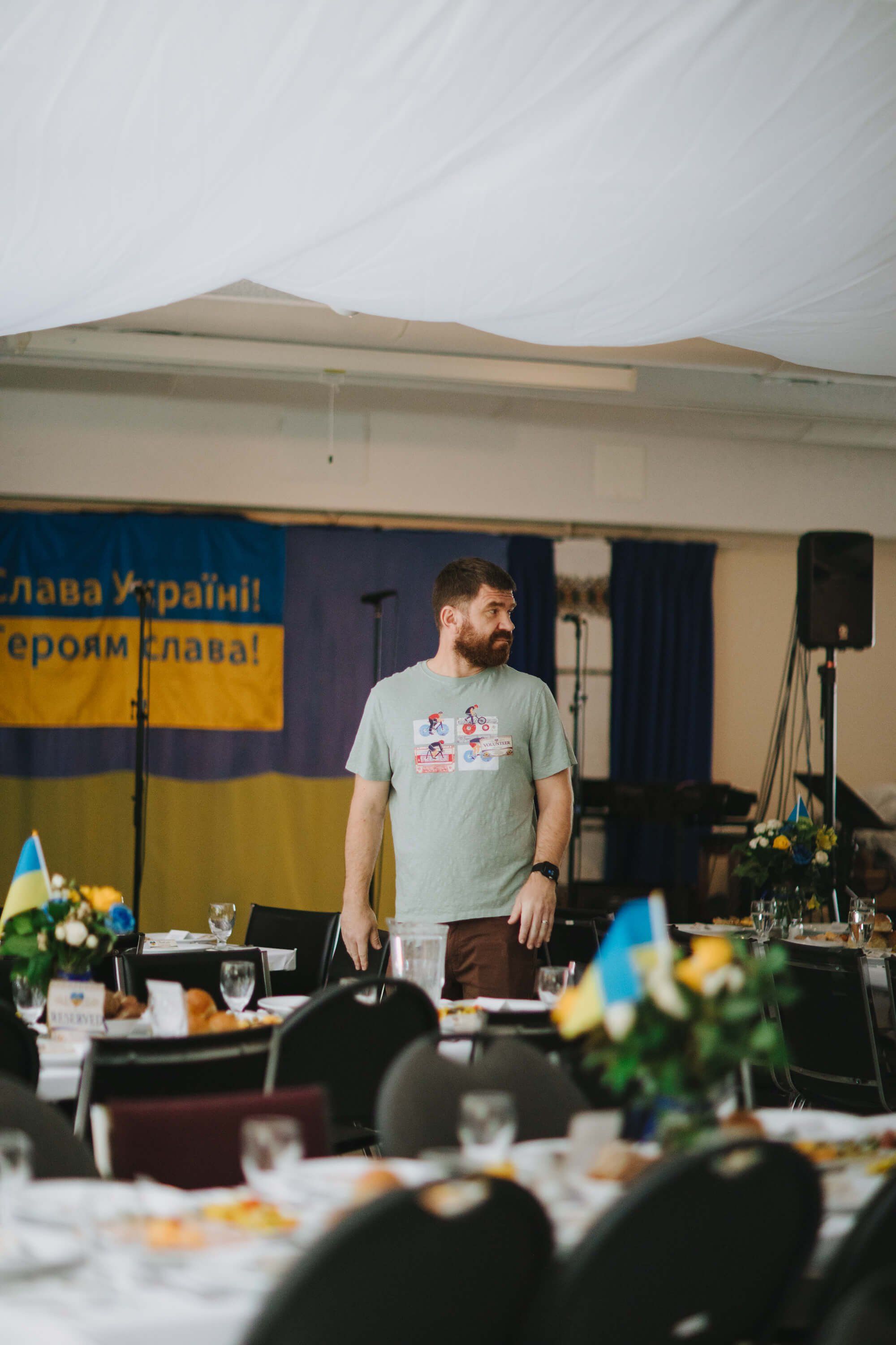 Man in light blue shirt stands at banquet table with Ukrainian flag backdrop and floral centerpieces