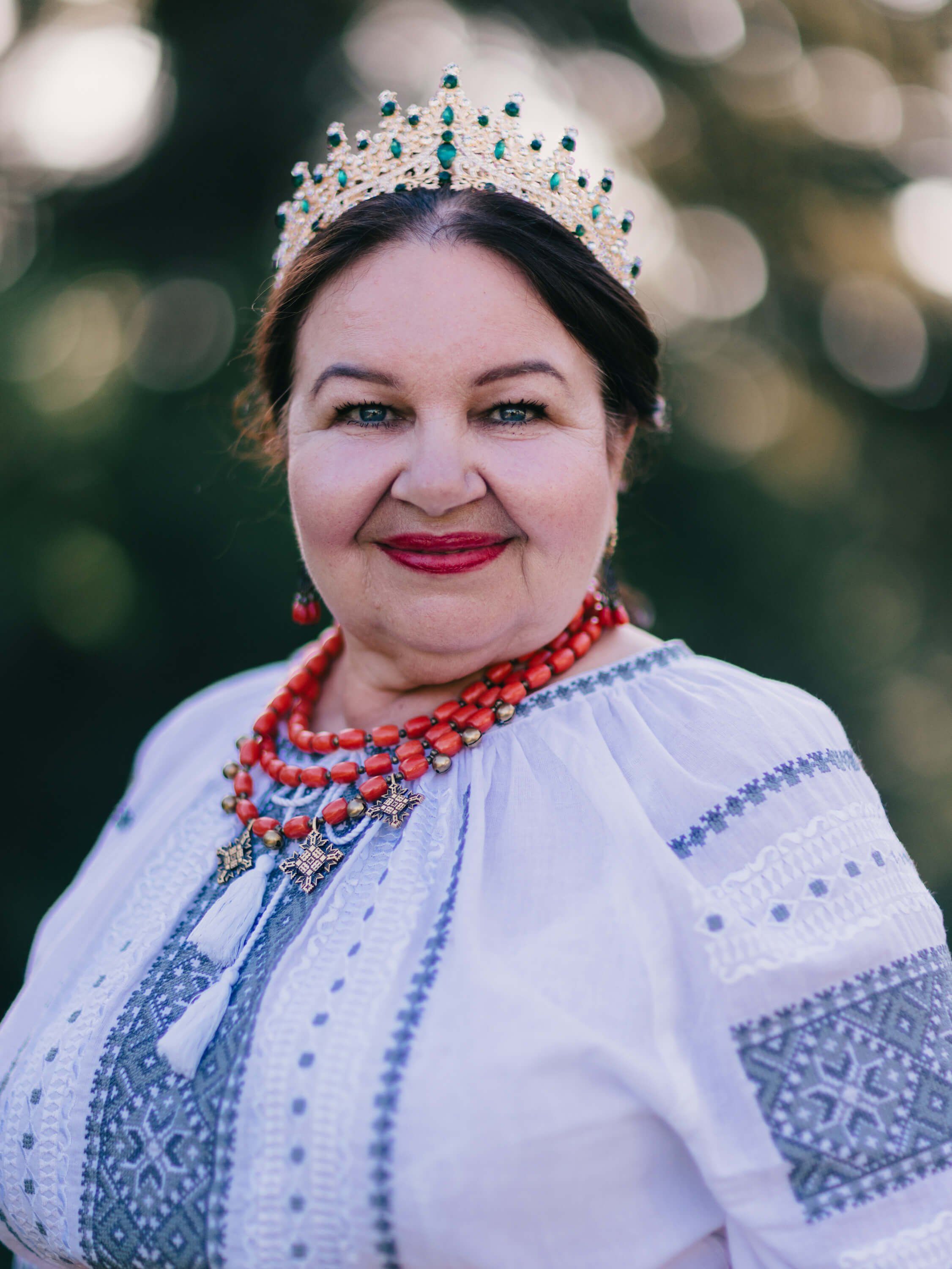 Woman in traditional Ukrainian embroidered white dress and beaded coral necklace, wearing an ornate tiara, smiles warmly against a softly blurred natural bac…
