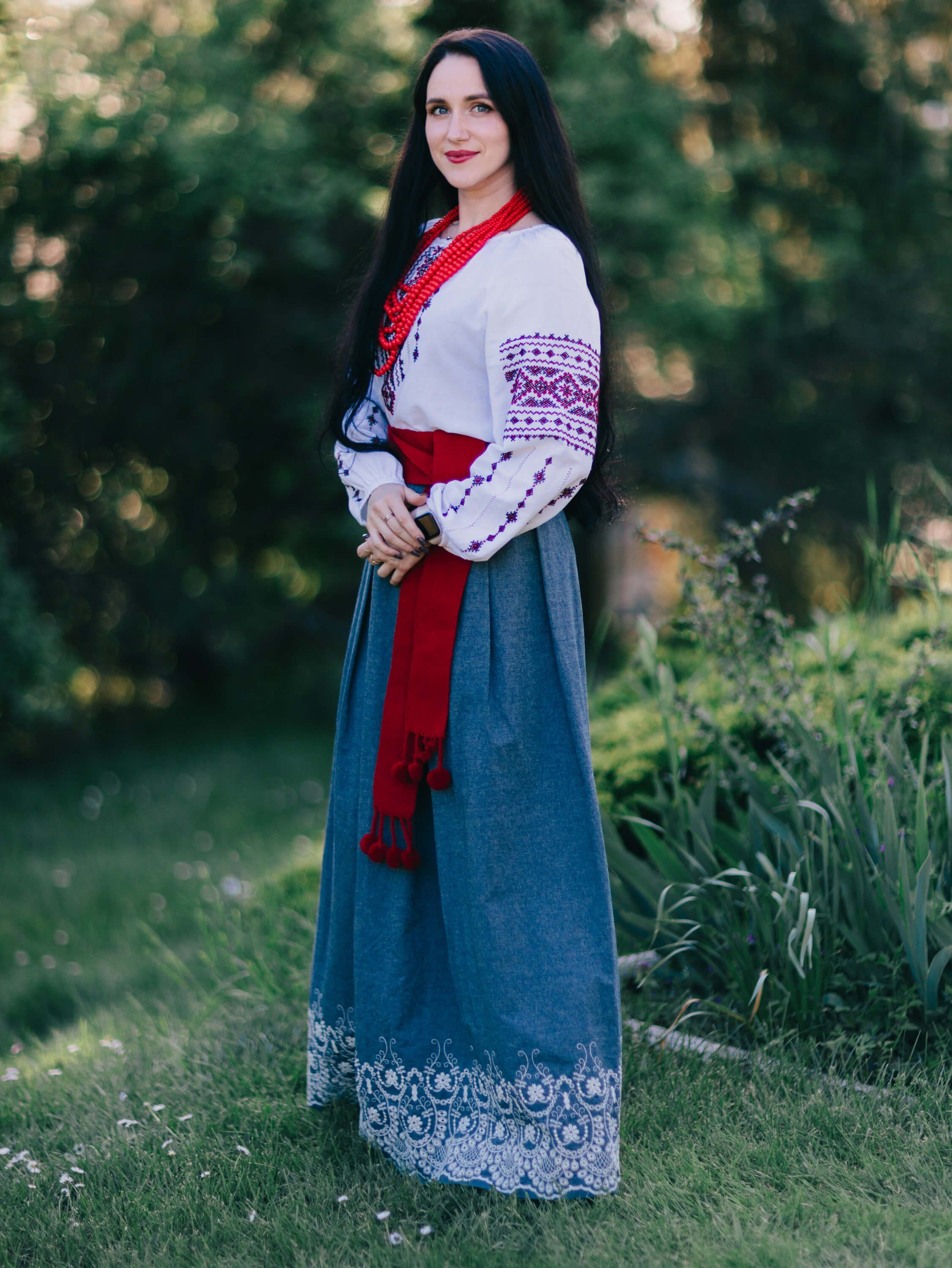 Woman in traditional Ukrainian embroidered dress and red sash stands in golden-hour garden light with evergreen trees behind her
