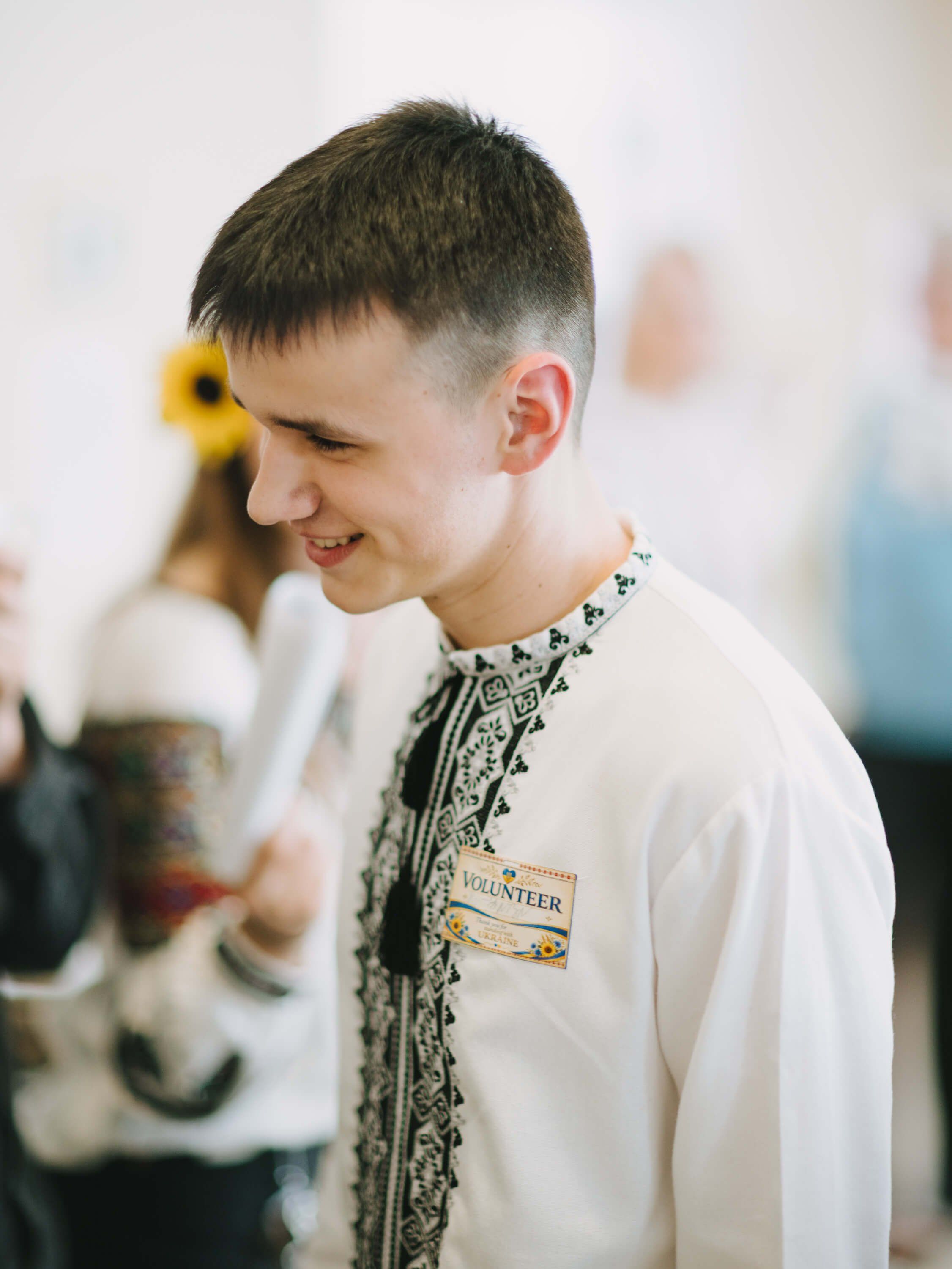Young man in cream embroidered shirt smiling at Ukrainian fundraiser dinner, soft indoor lighting