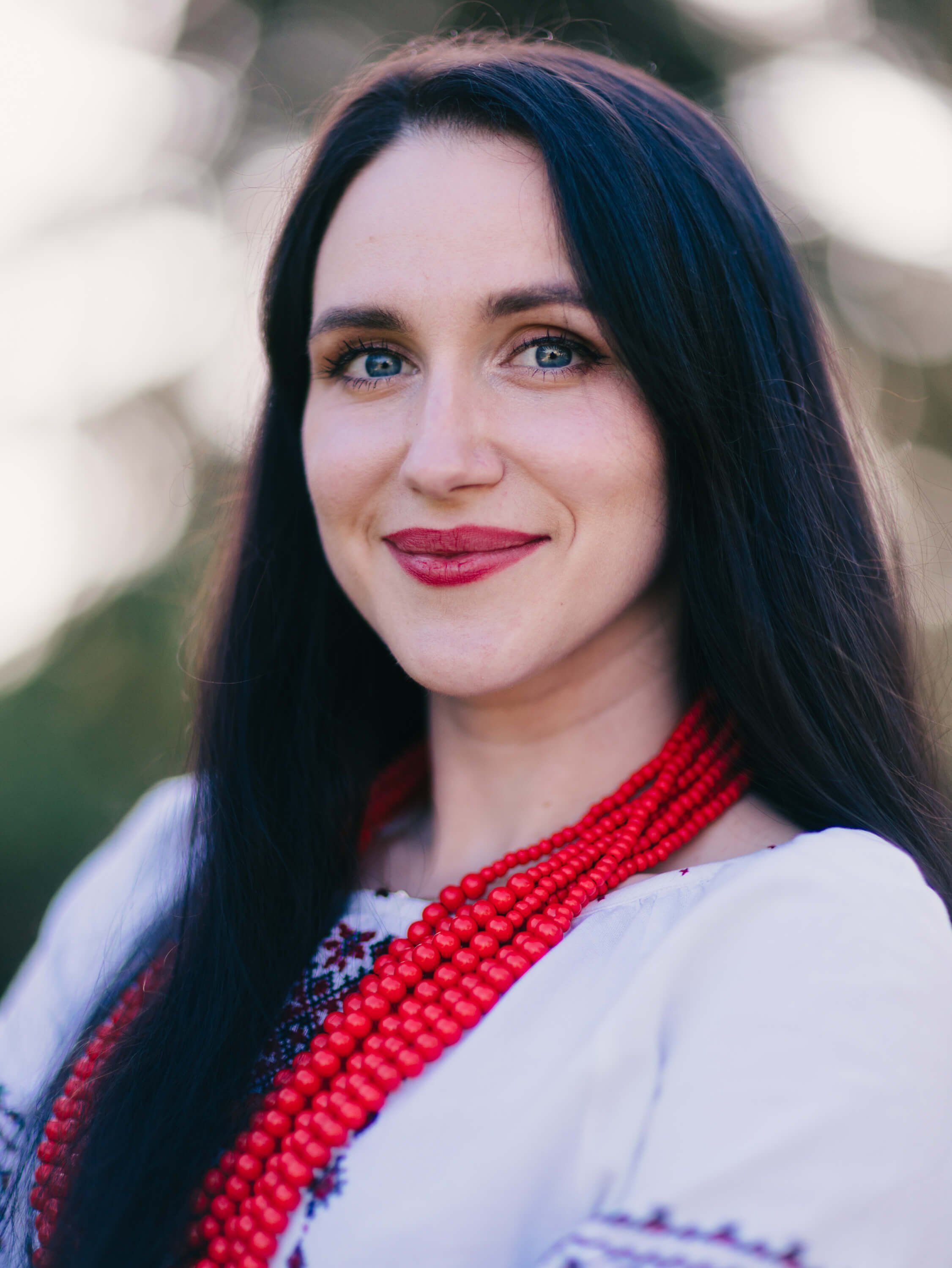 Woman with long dark hair and red lipstick wearing embroidered traditional Ukrainian clothing with red beaded necklace, outdoor portrait