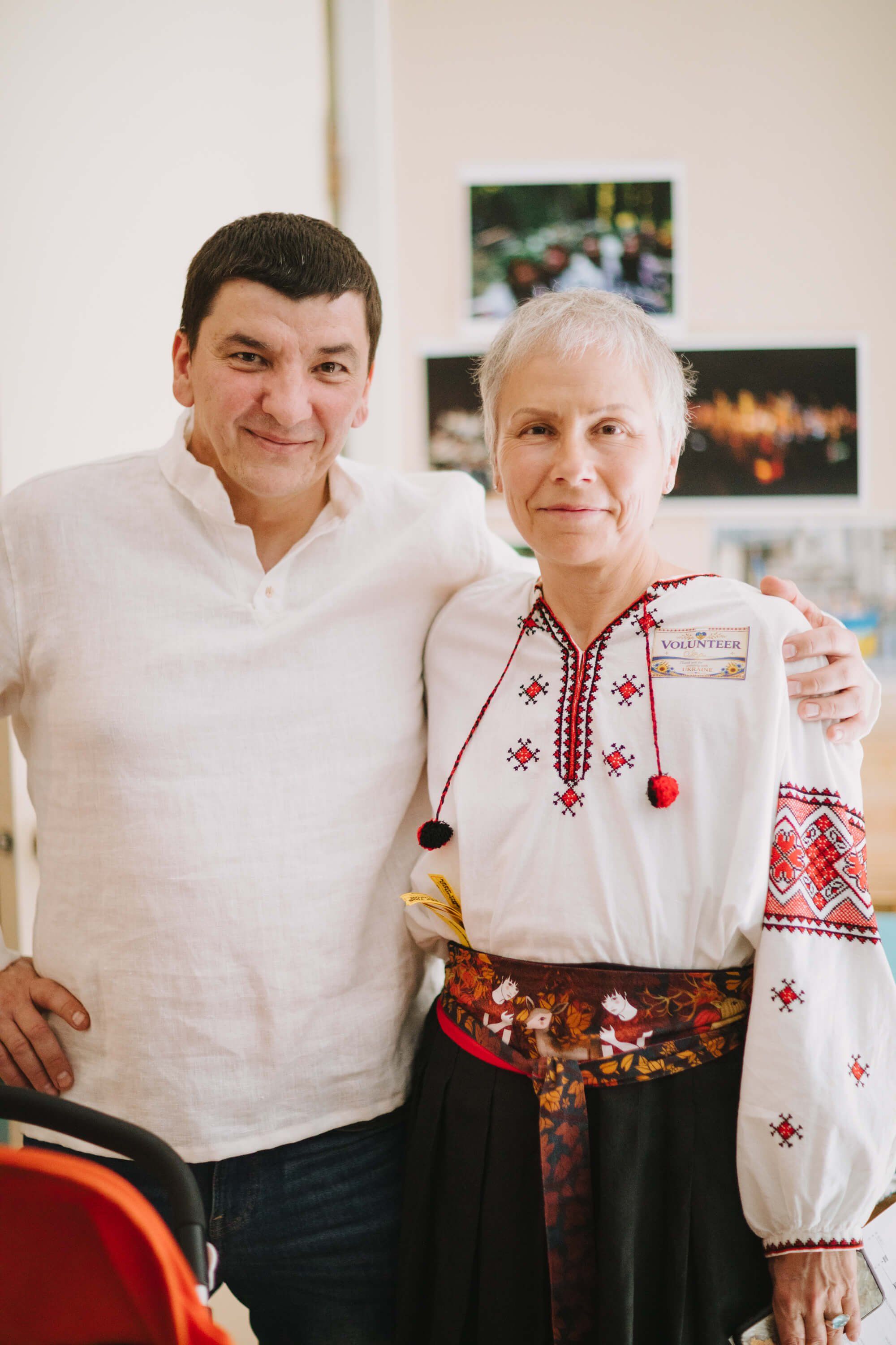 Two people embrace warmly indoors, the woman wearing traditional Ukrainian embroidered clothing with red geometric patterns and a dark skirt, the man in a wh…