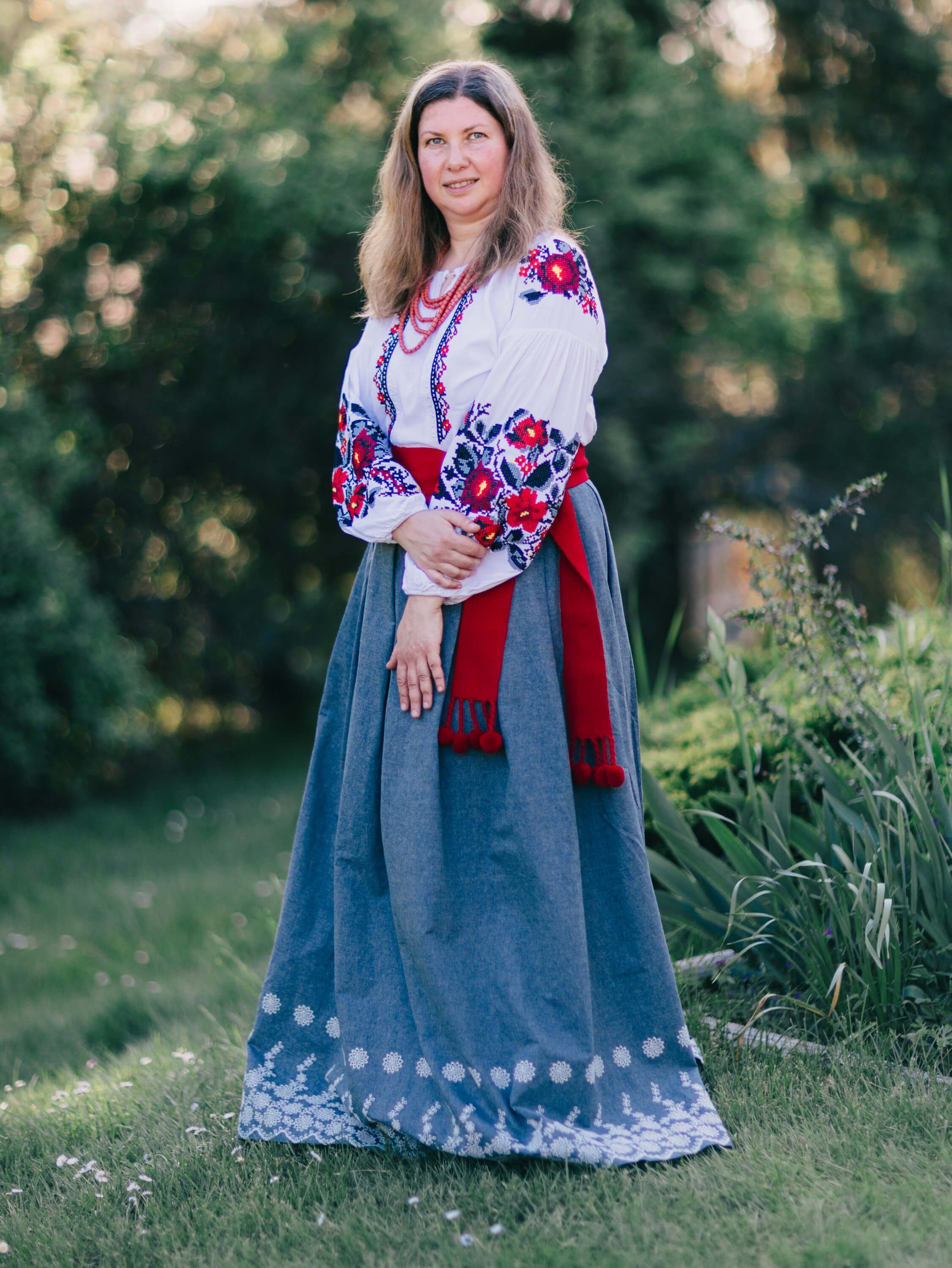 Woman in traditional Ukrainian embroidered blouse and blue skirt with lace hem stands in golden-hour garden light