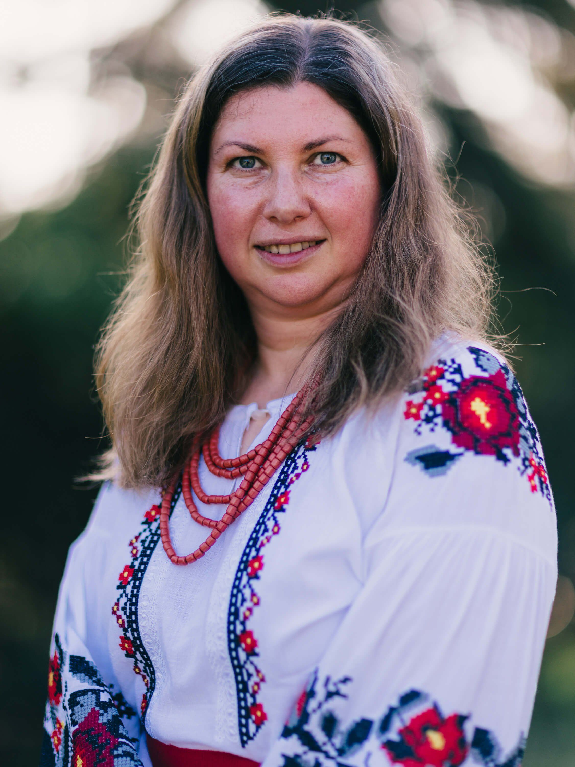 Woman in white embroidered Ukrainian blouse with red beaded necklaces smiles at camera outdoors at golden hour
