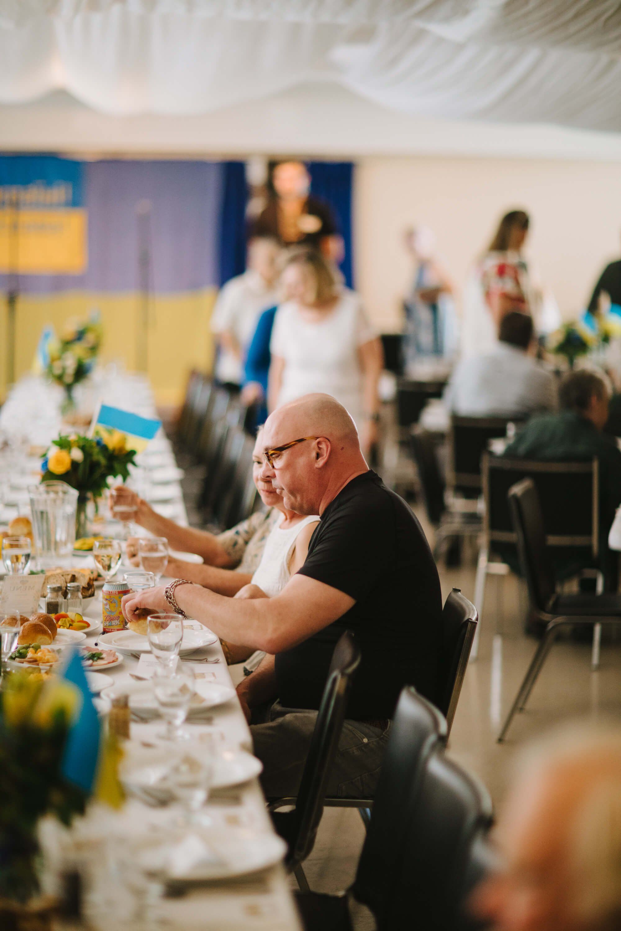 Man in glasses and black shirt gestures while seated at a long table during a Ukrainian fundraiser dinner, with guests mingling in the background