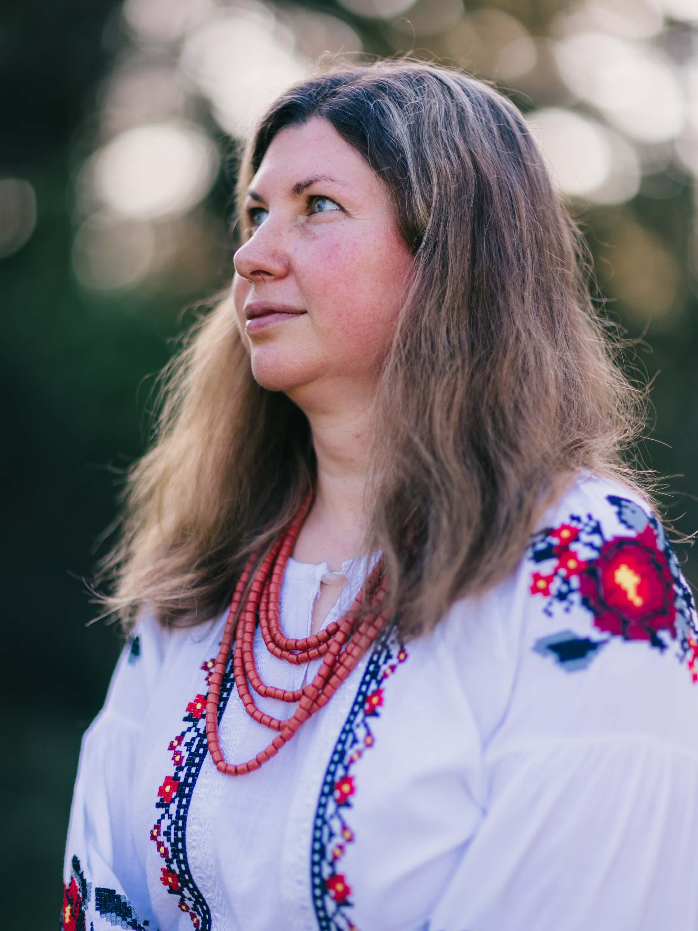 Woman in traditional Ukrainian embroidered white linen shirt with red beading gazes thoughtfully into distance against soft-focused garden backdrop