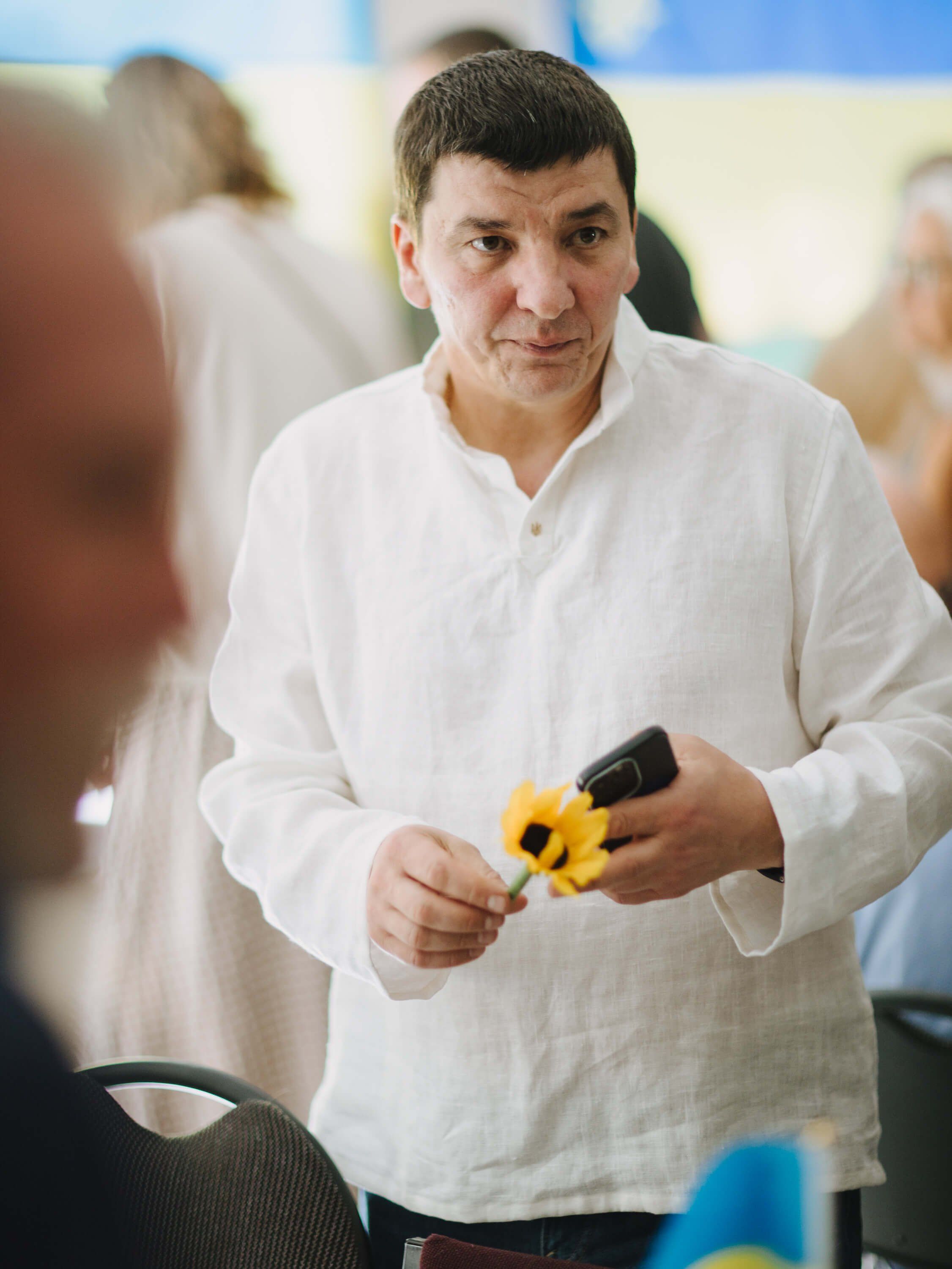 Man in white shirt holding yellow tools speaks to attendees at a Ukrainian fundraiser dinner event indoors