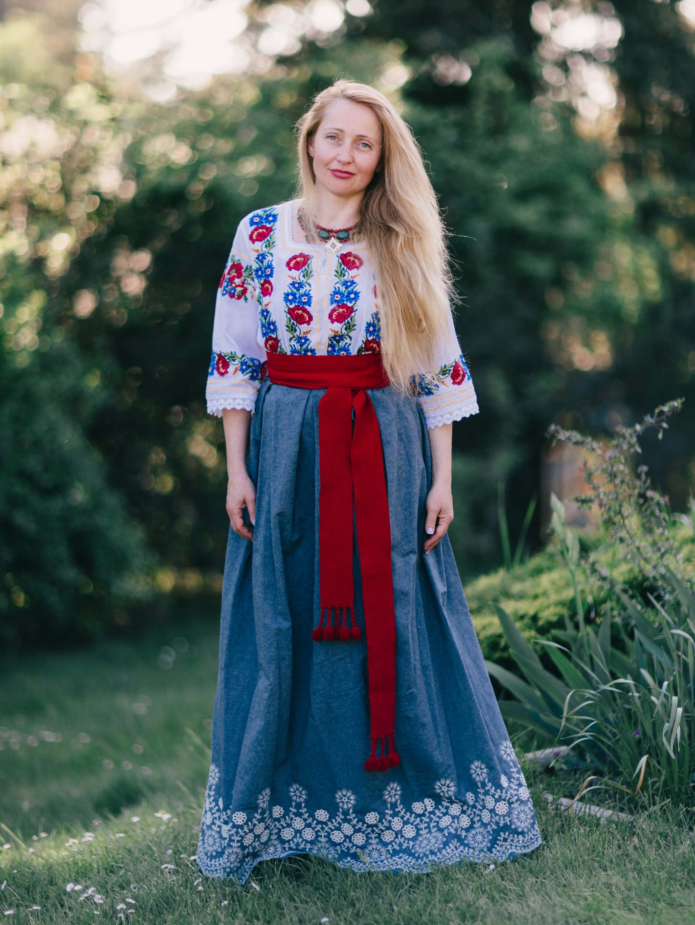 Woman in embroidered white blouse and denim skirt with red sash standing in golden-hour garden light, soft-focused greenery behind