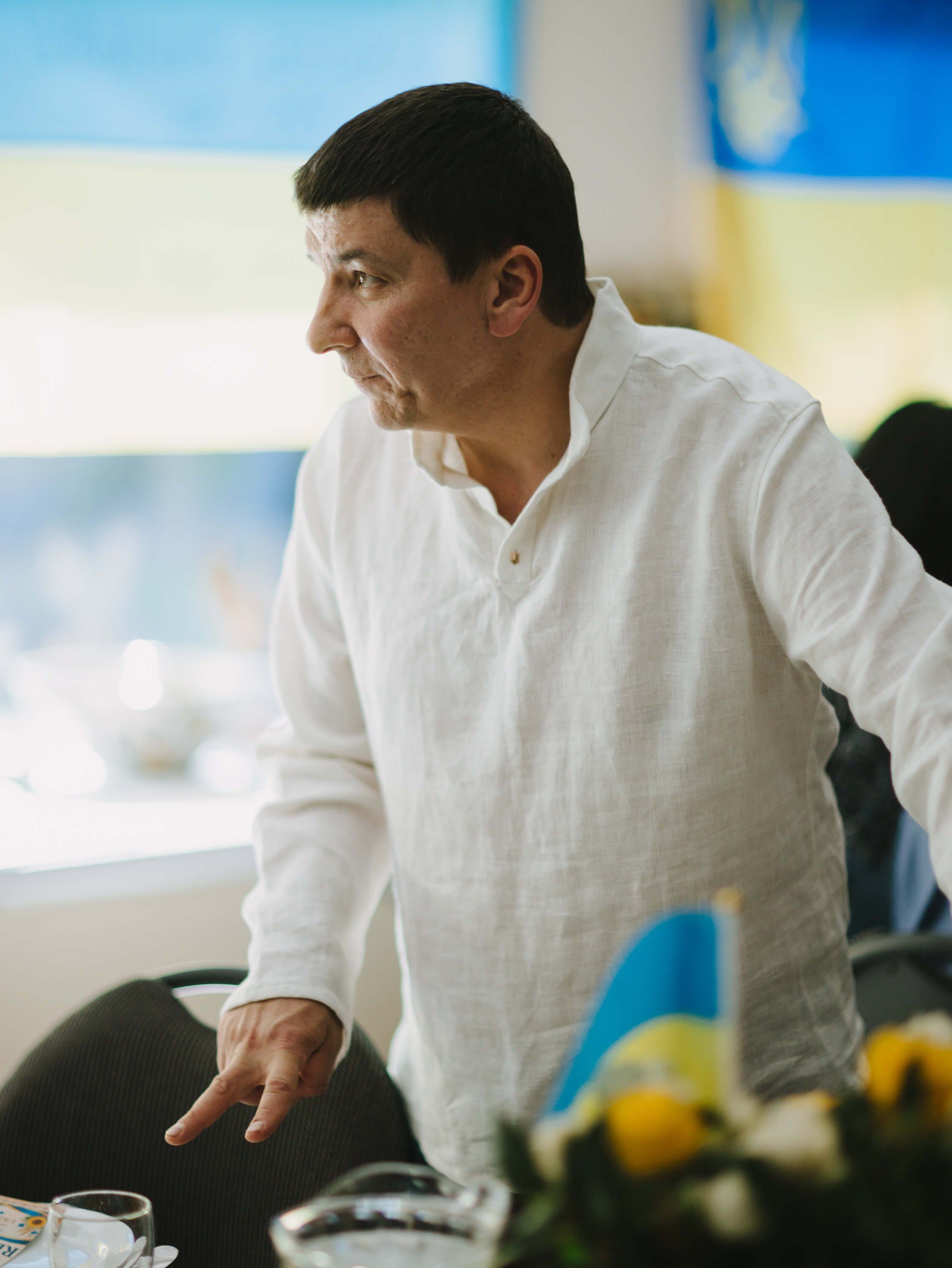 Man in white shirt speaking animatedly at a Ukrainian fundraiser dinner, with blue and yellow decor visible behind him