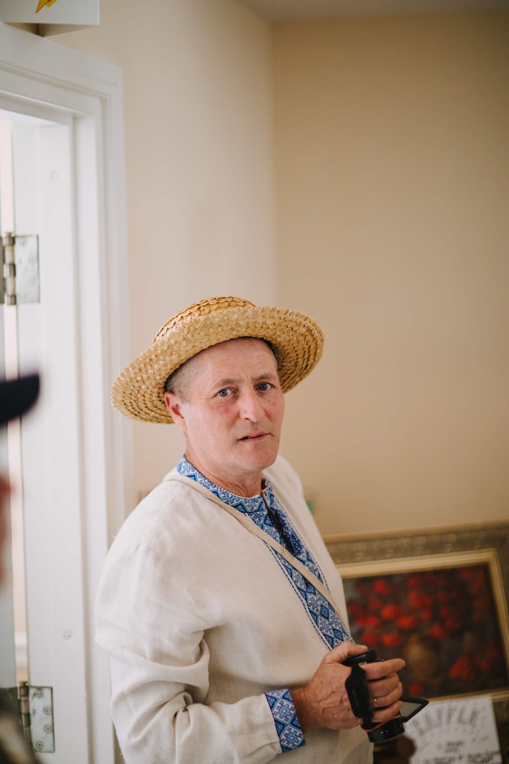 Man in cream linen shirt and straw hat with blue embroidered collar turns toward camera in warm-lit interior space