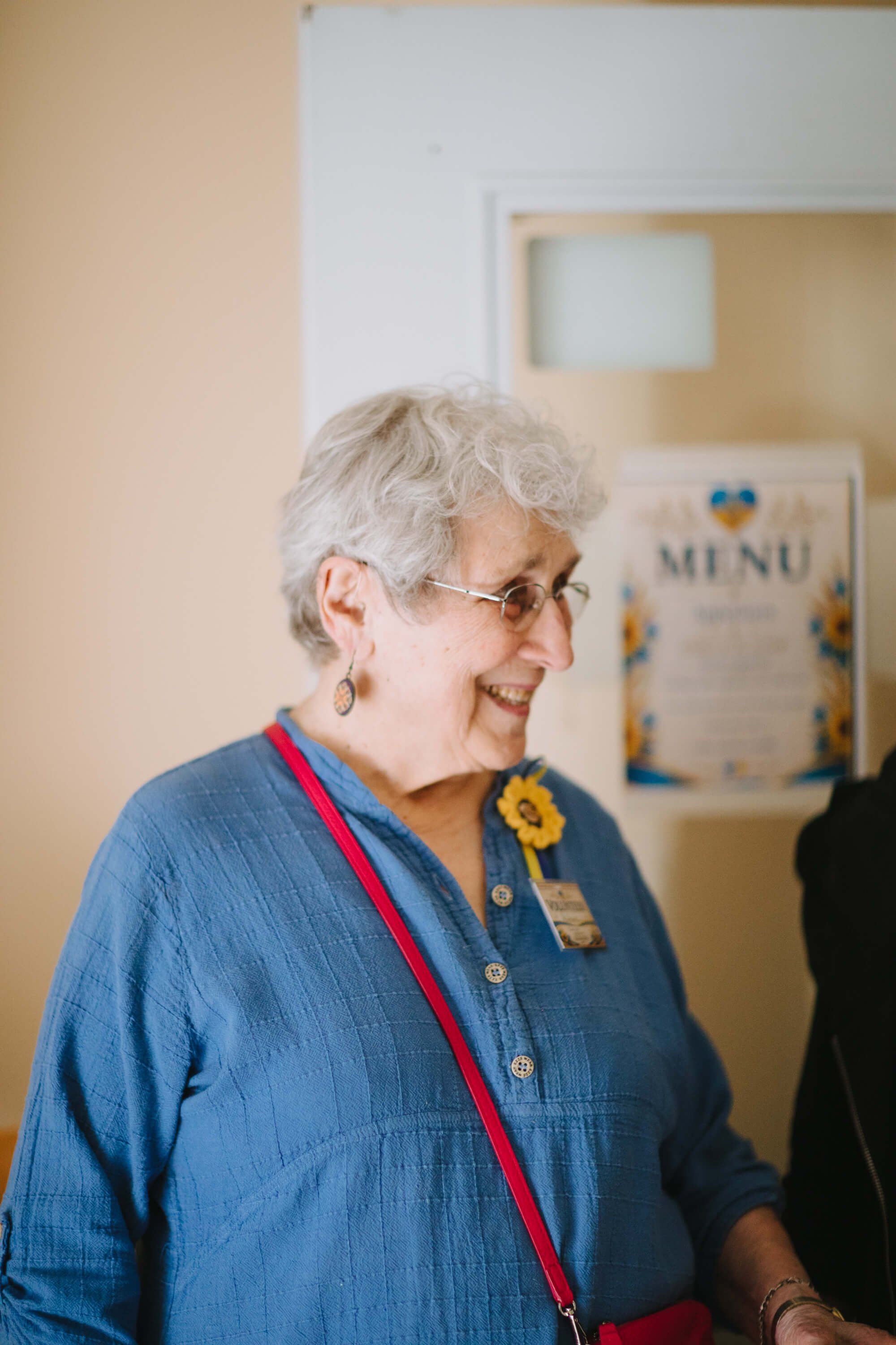 Older woman with white hair wearing glasses and a blue shirt with a sunflower pin and red lanyard smiles at a community event