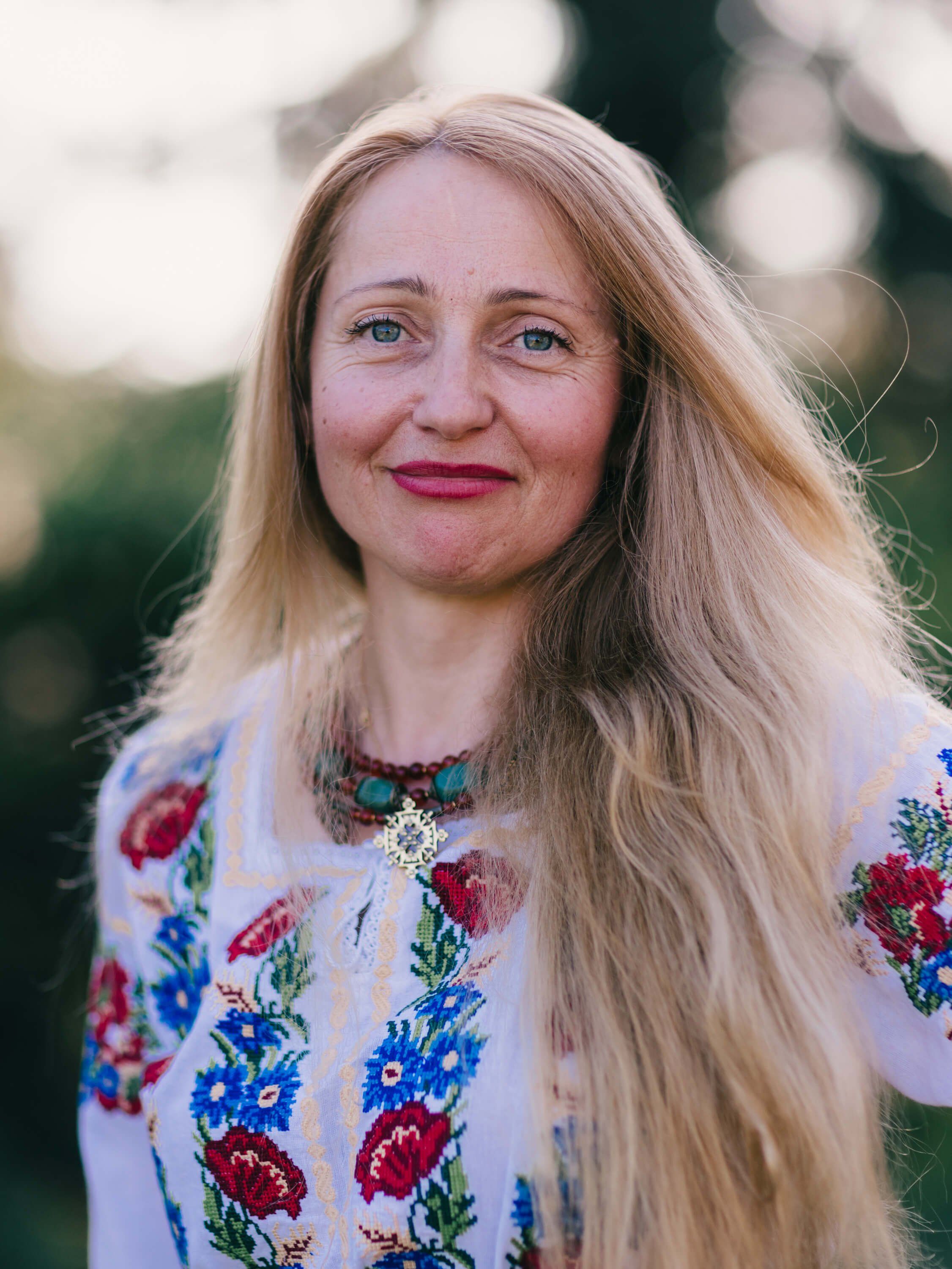 Woman with long blonde hair wearing an embroidered white blouse with red and blue floral patterns, smiling outdoors in soft natural light