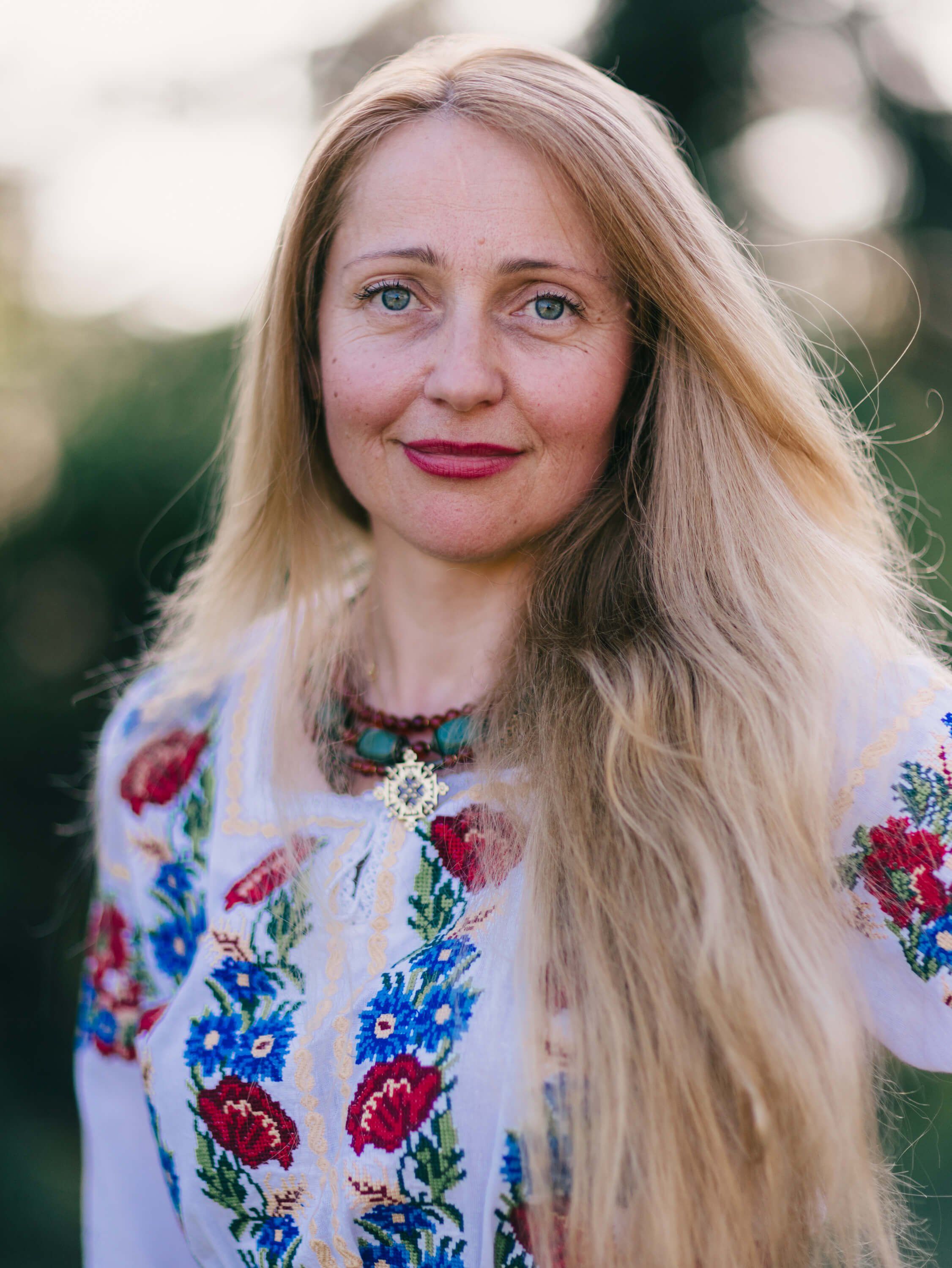 Woman with long blonde hair wearing an embroidered white blouse with red and blue floral details, outdoors with soft natural light
