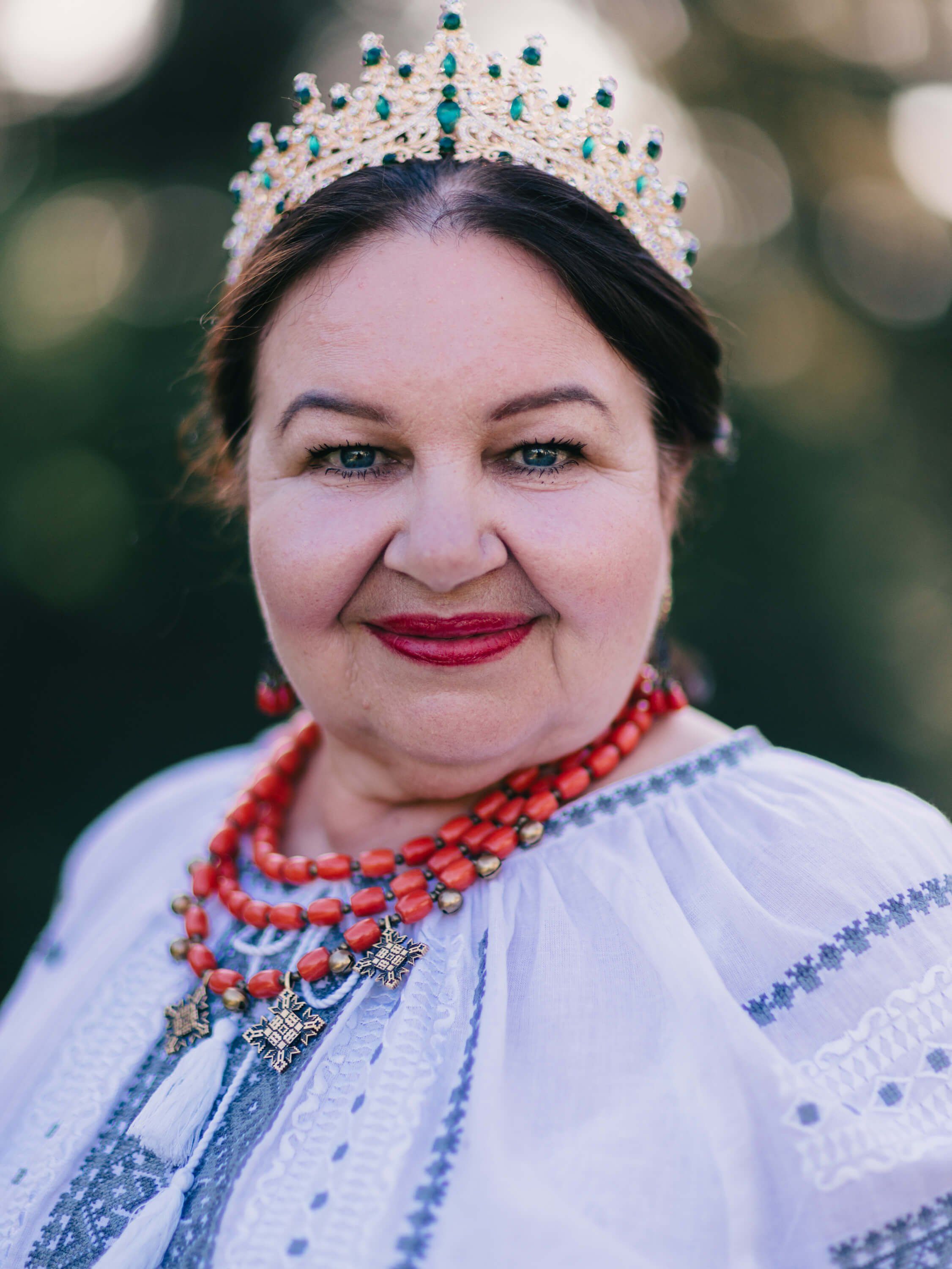 Woman in traditional Ukrainian embroidered white dress and beaded tiara smiles warmly at camera, coral beads glowing against soft blurred background