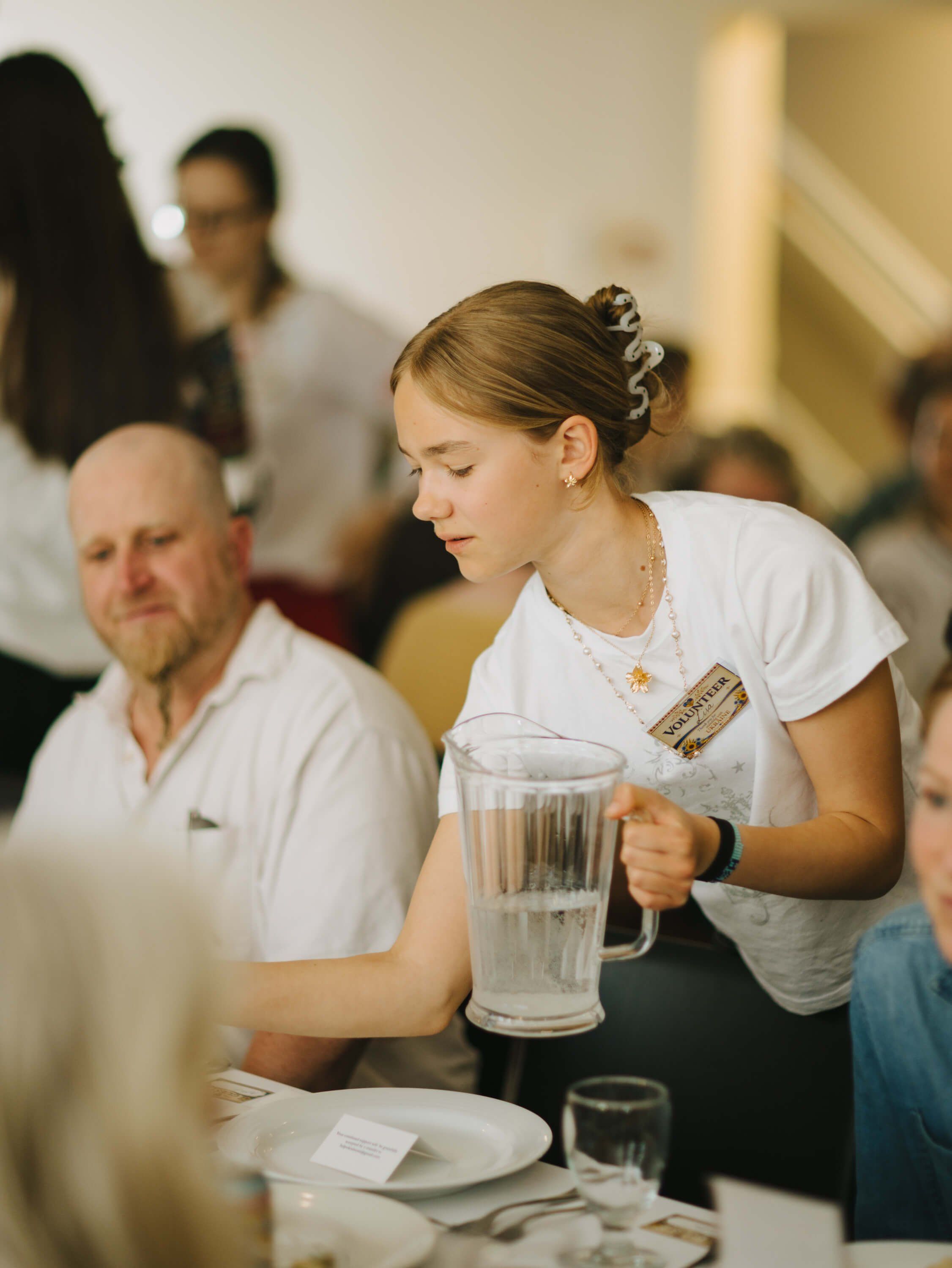 Young woman in white shirt pouring water at a candlelit dinner table surrounded by guests in warm indoor lighting