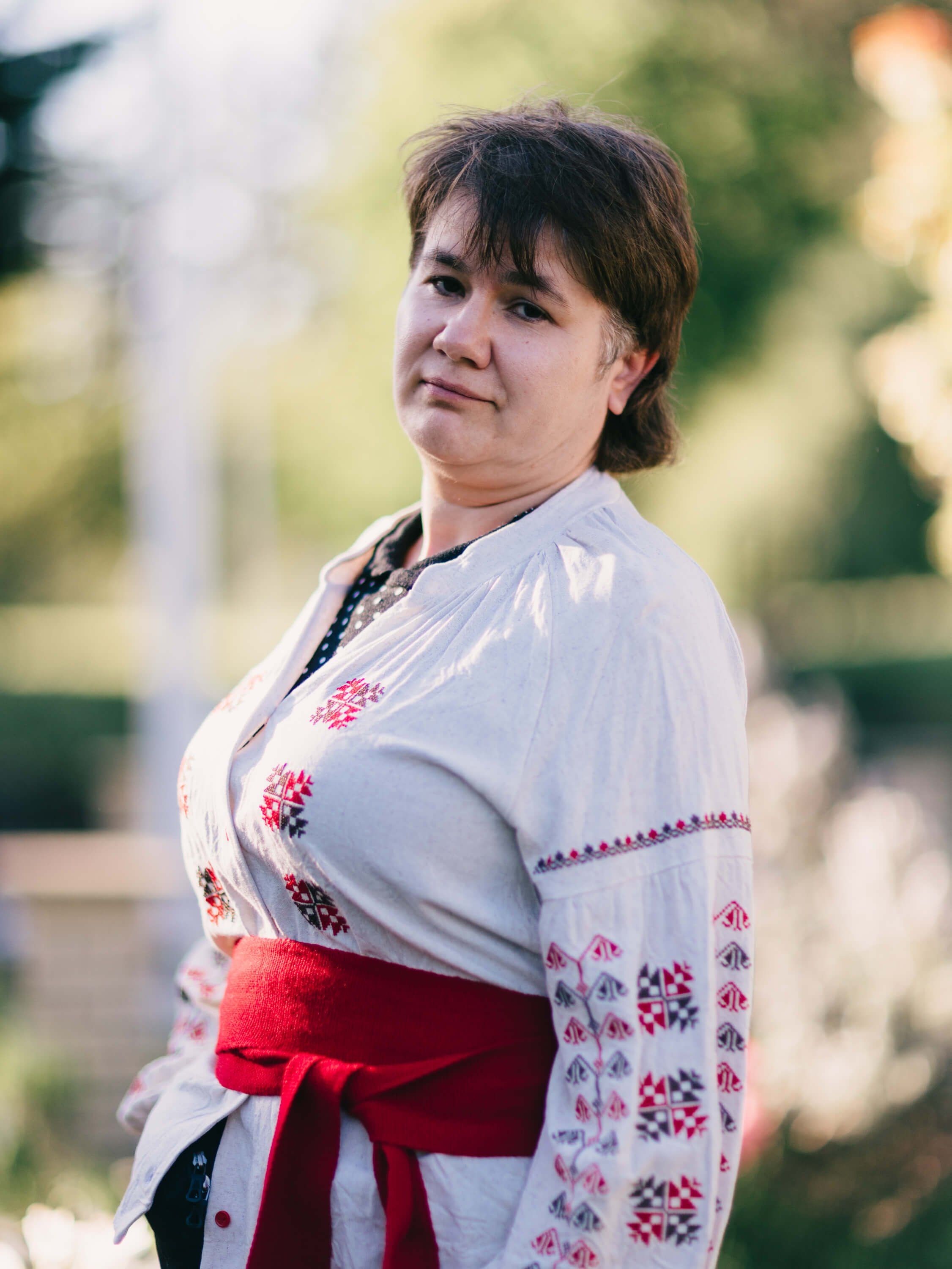 Woman in embroidered Ukrainian blouse with red sash smiles at camera in soft outdoor light