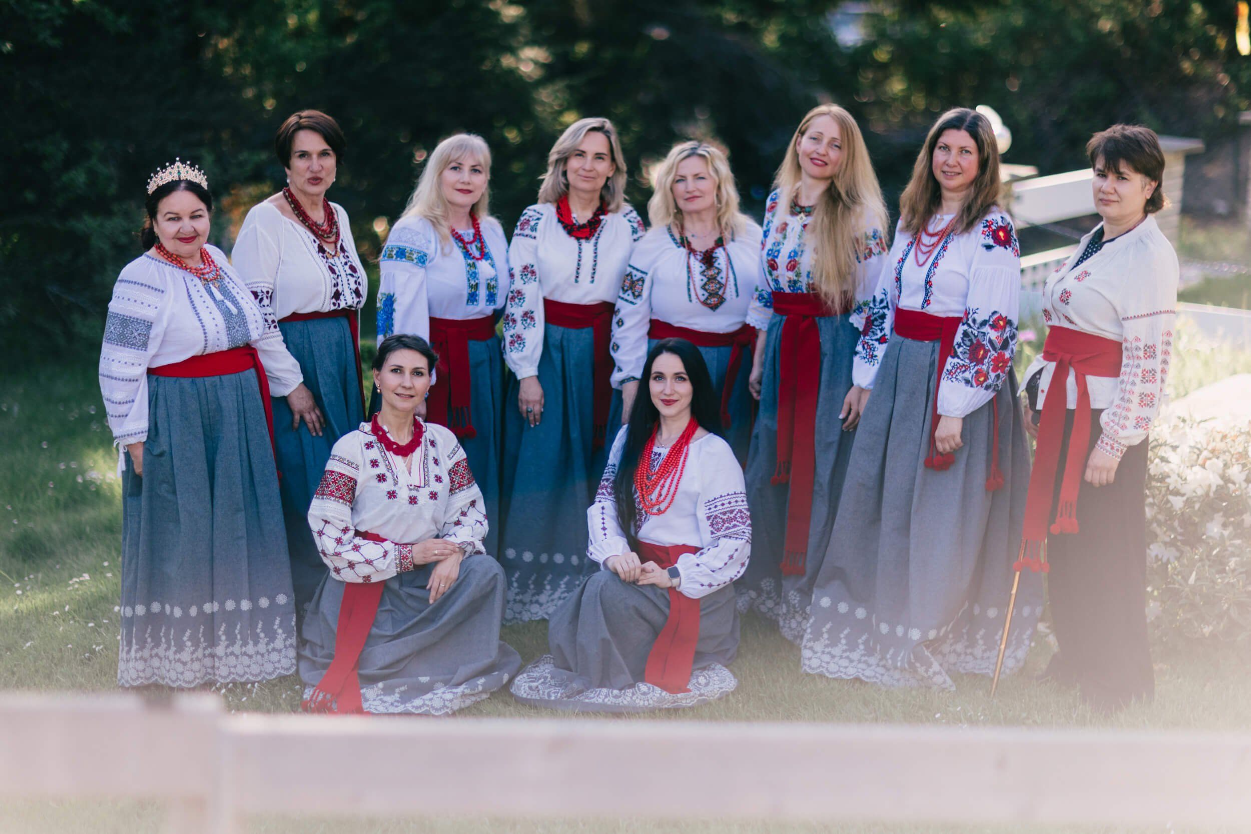 Ten women in traditional Ukrainian embroidered blouses and colorful skirts pose together outdoors in warm, golden light