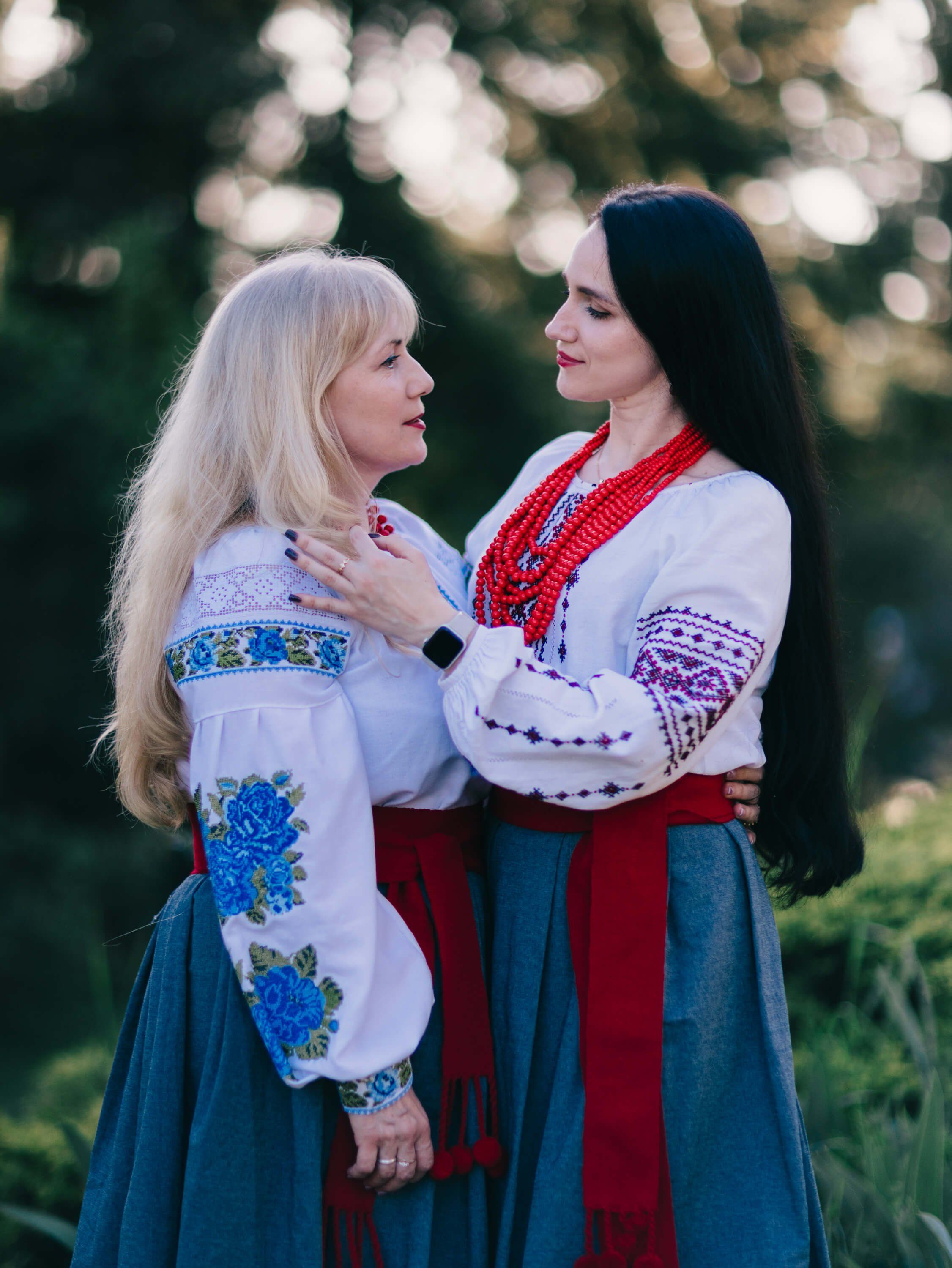 Two women in embroidered Ukrainian dress embrace outdoors at golden hour, their traditional vyshyvankas adorned with blue and red geometric patterns