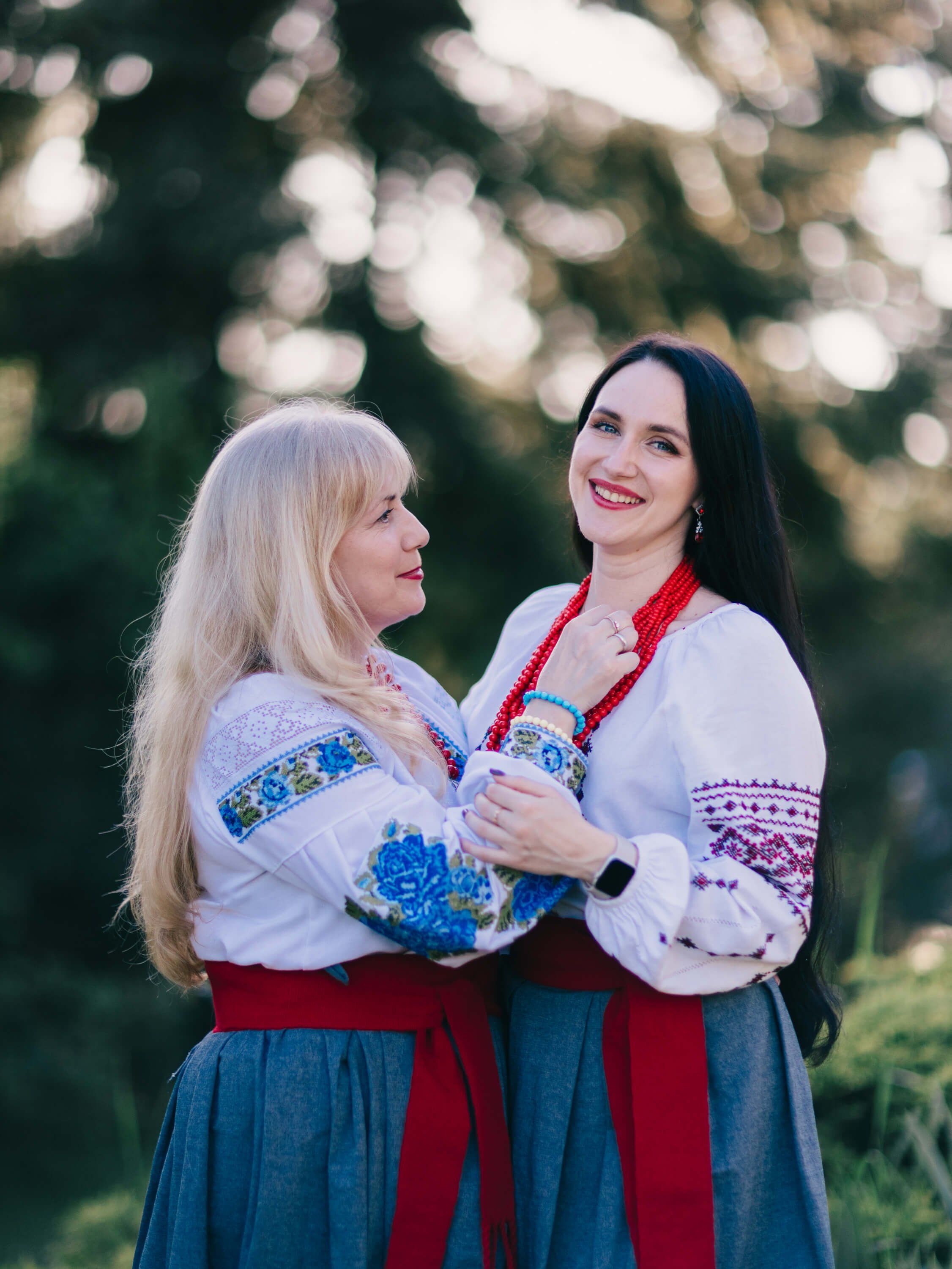 Two women in traditional Ukrainian embroidered blouses smile warmly at each other outdoors at golden hour, with soft bokeh foliage glowing behind them
