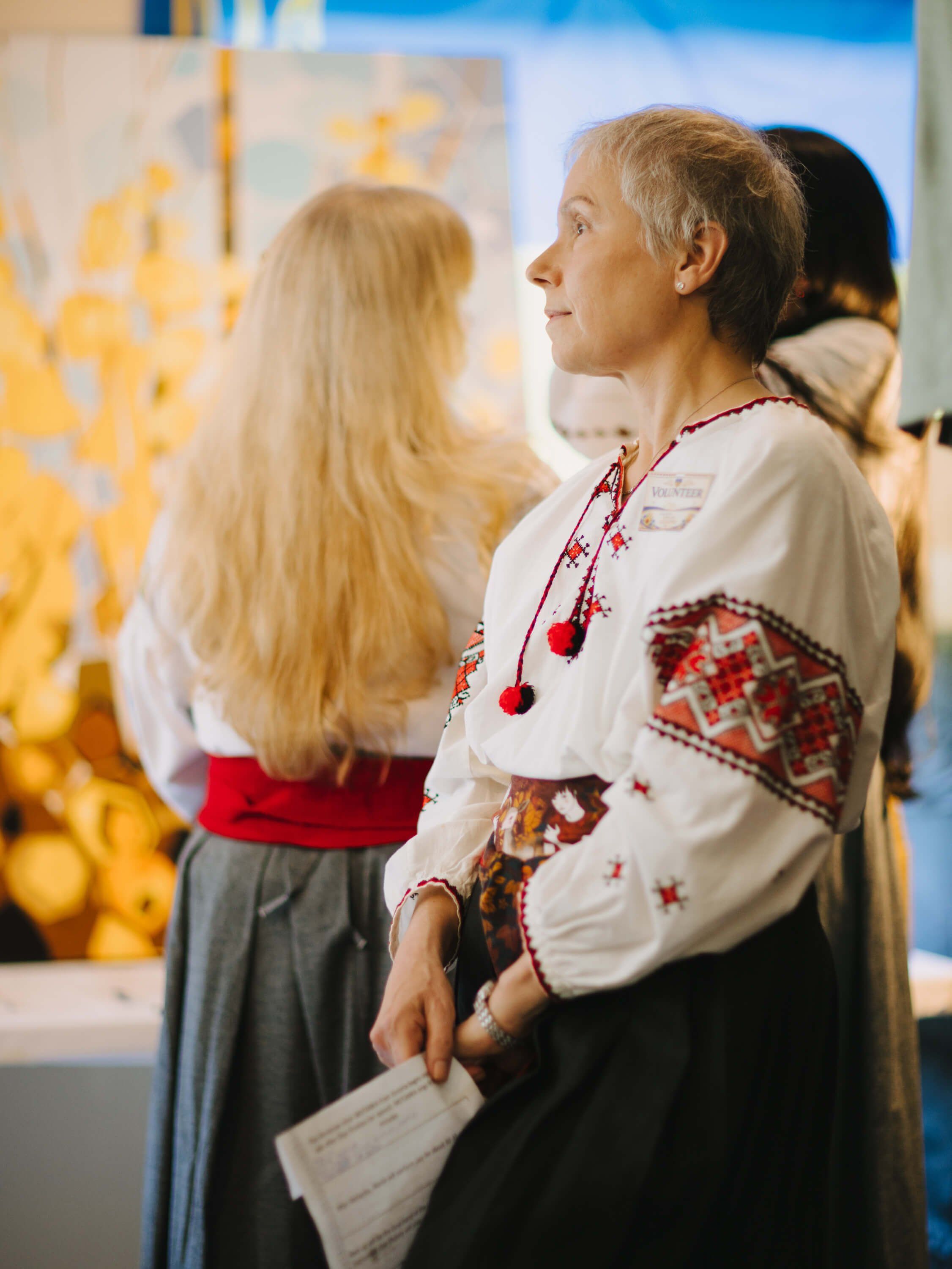 Elegant guest in embroidered Ukrainian blouse gazes at golden artwork during evening fundraiser event