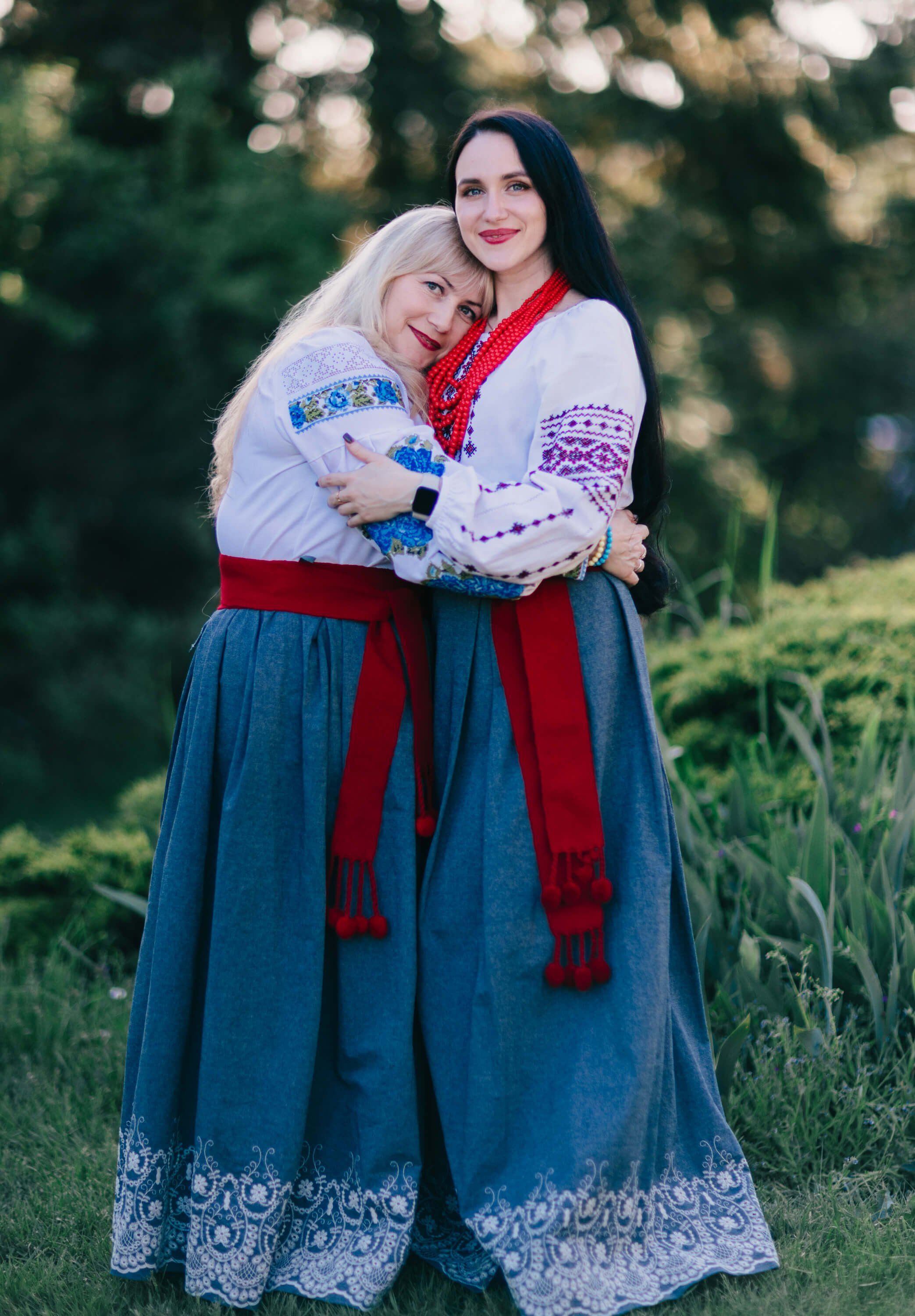 Two women in embroidered Ukrainian traditional dress embrace outdoors at golden hour, surrounded by soft green foliage