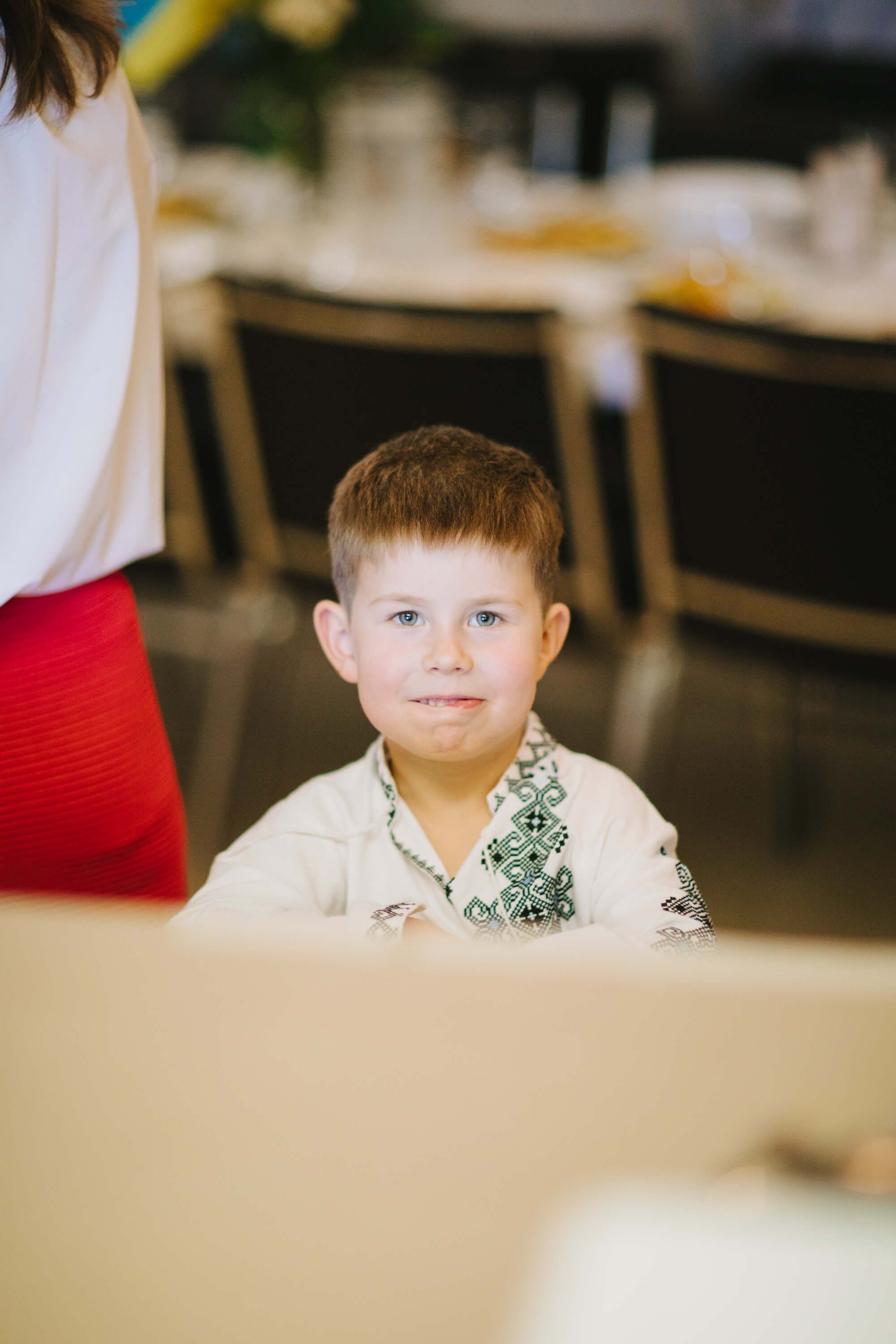 Young boy in green patterned bow tie sits at formal dinner table, gazing thoughtfully at camera