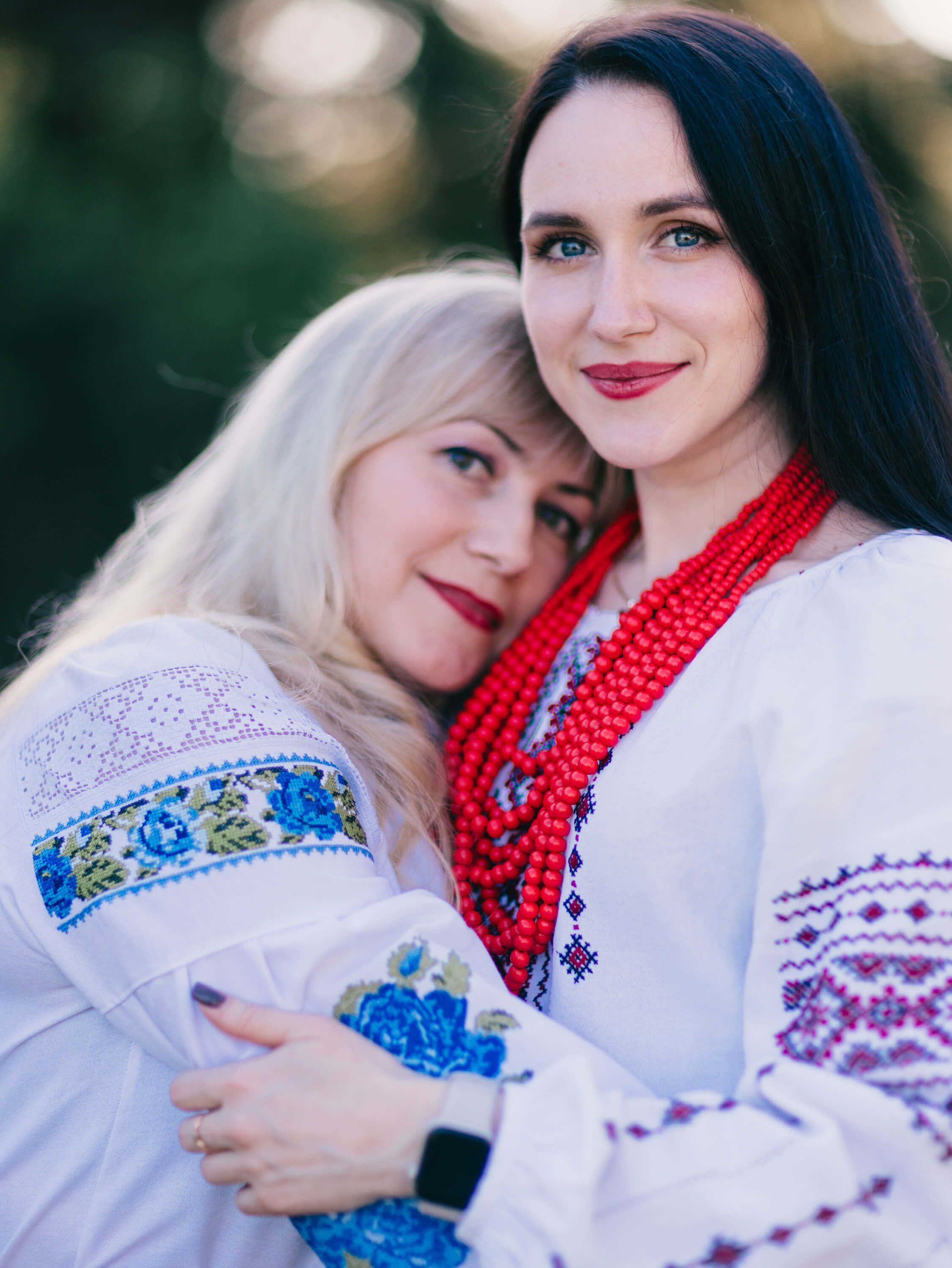 Two women in embroidered white Ukrainian garments embrace outdoors at golden hour, their warm smiles and traditional dress radiant against soft, blurred gree…